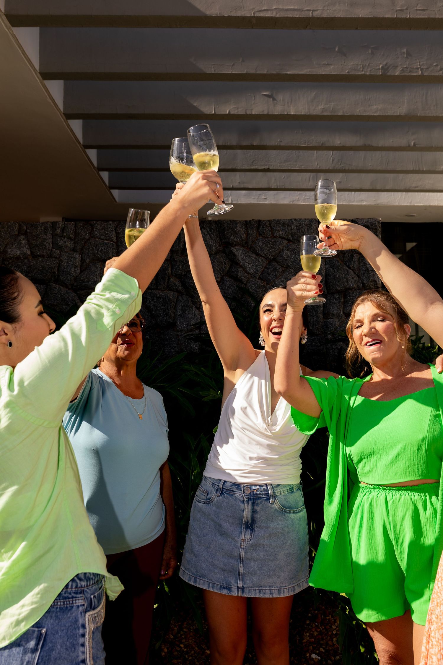 Four women raising glasses of white wine in a celebratory toast outdoors on a sunny day.
