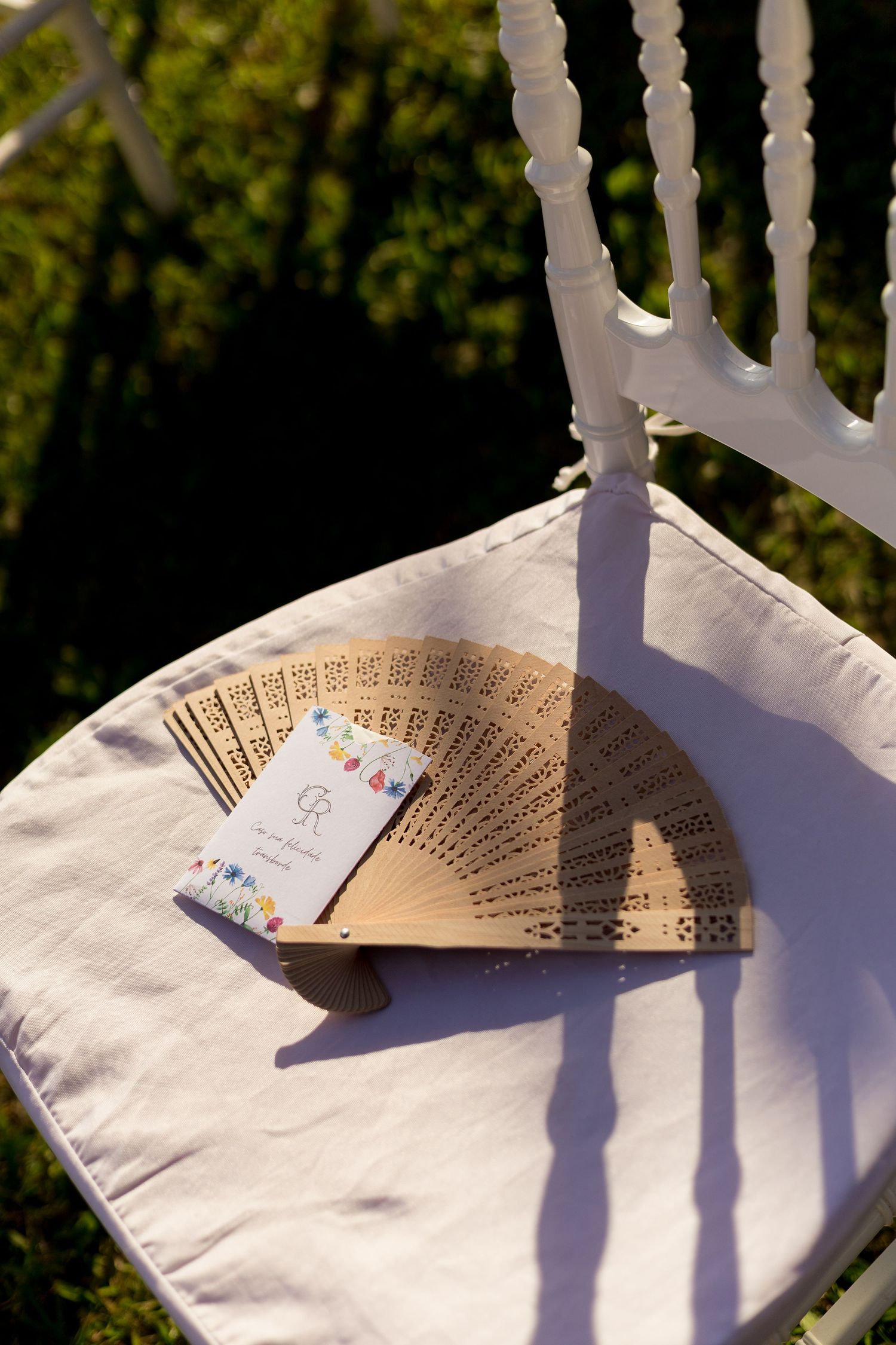 Wooden hand fan with decorative card on white chair cushion outdoors in sunlight.