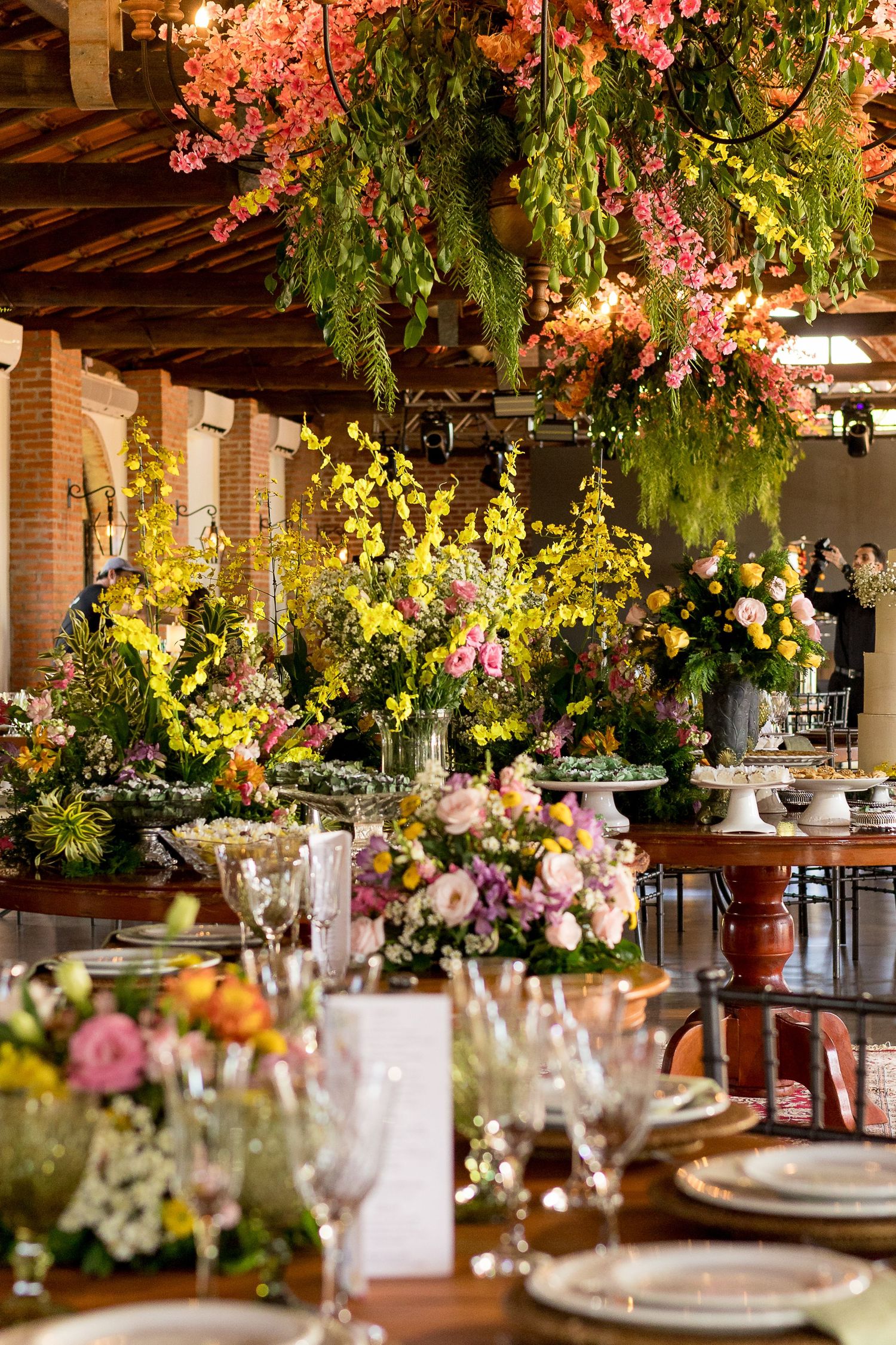 Elegant floral wedding reception with hanging greenery, yellow orchids, and colorful table arrangements in a rustic venue.