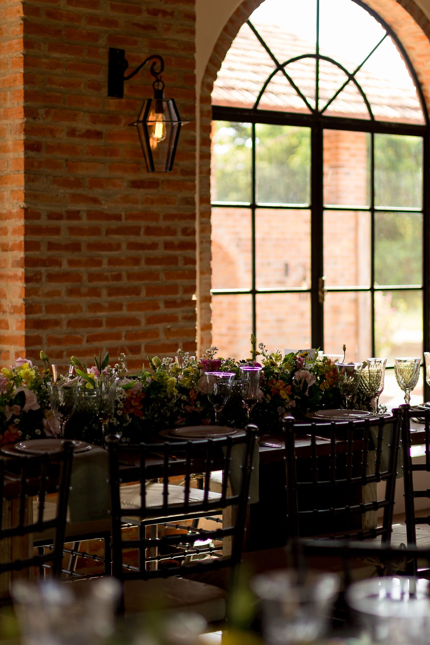 Elegant rustic wedding reception table with floral centerpieces, dark chairs, and arched windows in a brick venue.