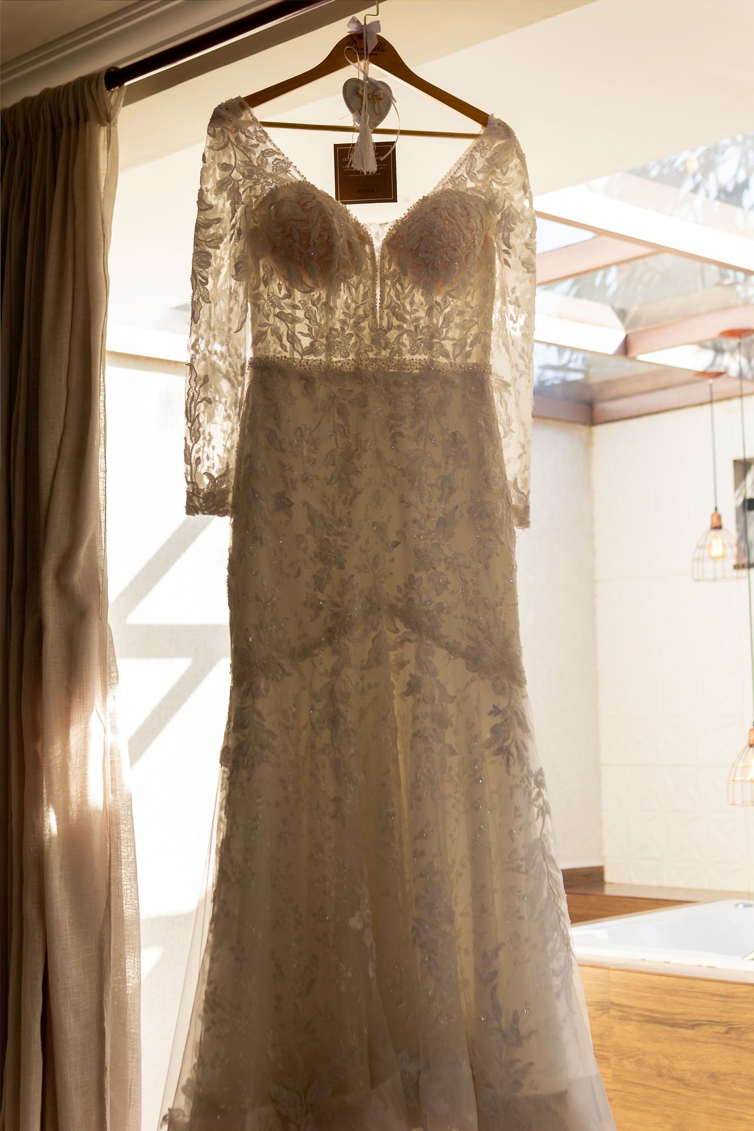 Elegant lace wedding dress with sheer sleeves hanging on a wooden hanger near a sunlit window.