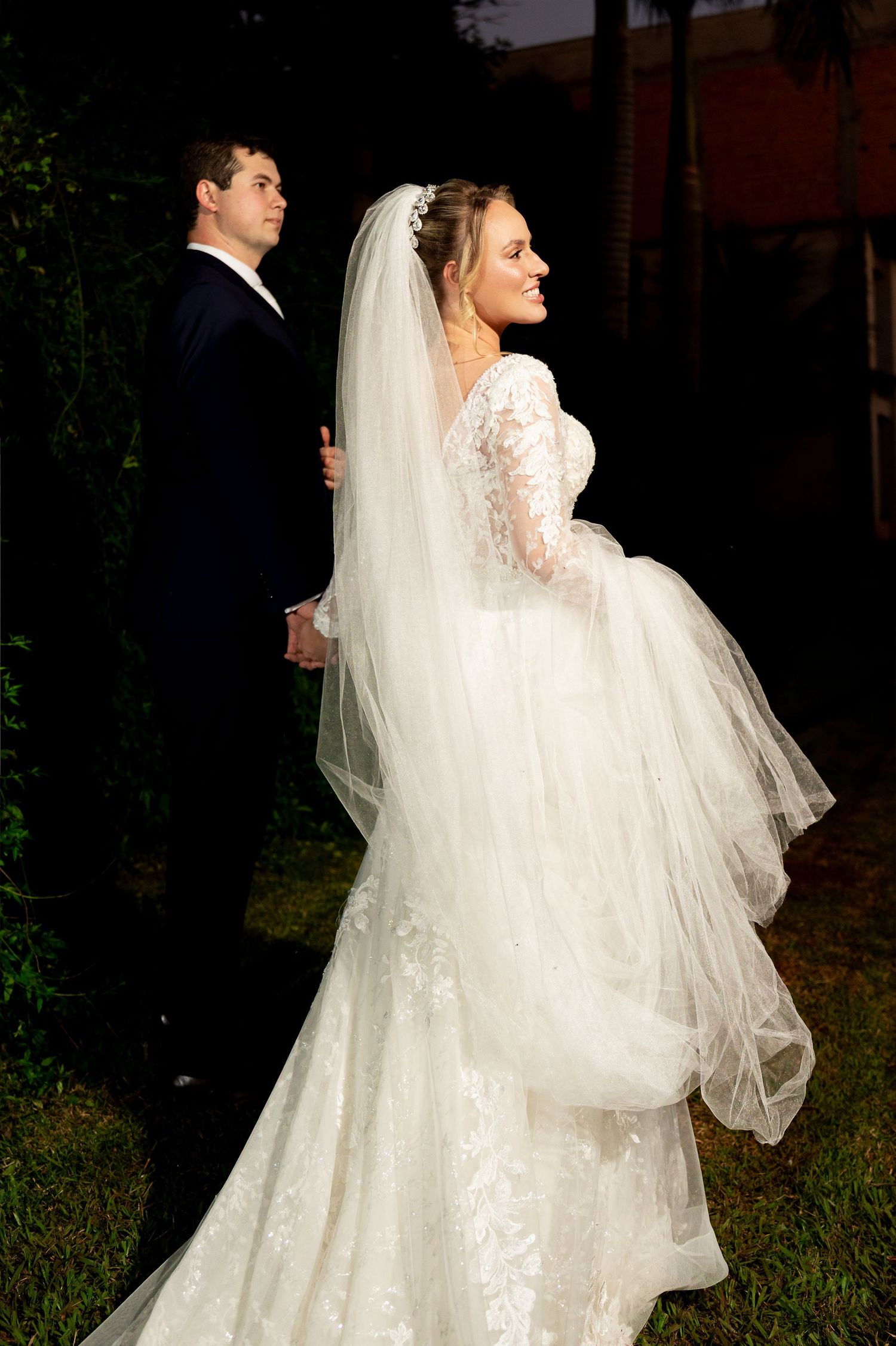 Bride in lace gown with veil holds groom's hand outdoors at night during elegant wedding portrait.