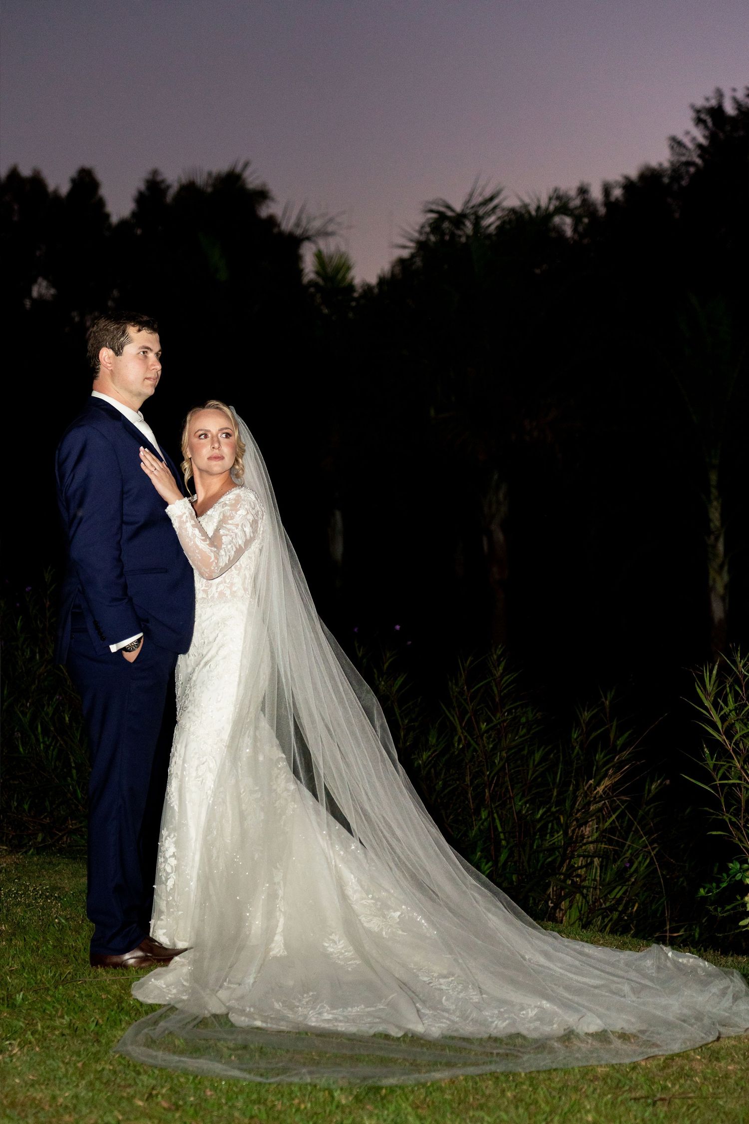 Bride in lace gown with cathedral veil stands with groom in navy suit at twilight outdoor wedding portrait.
