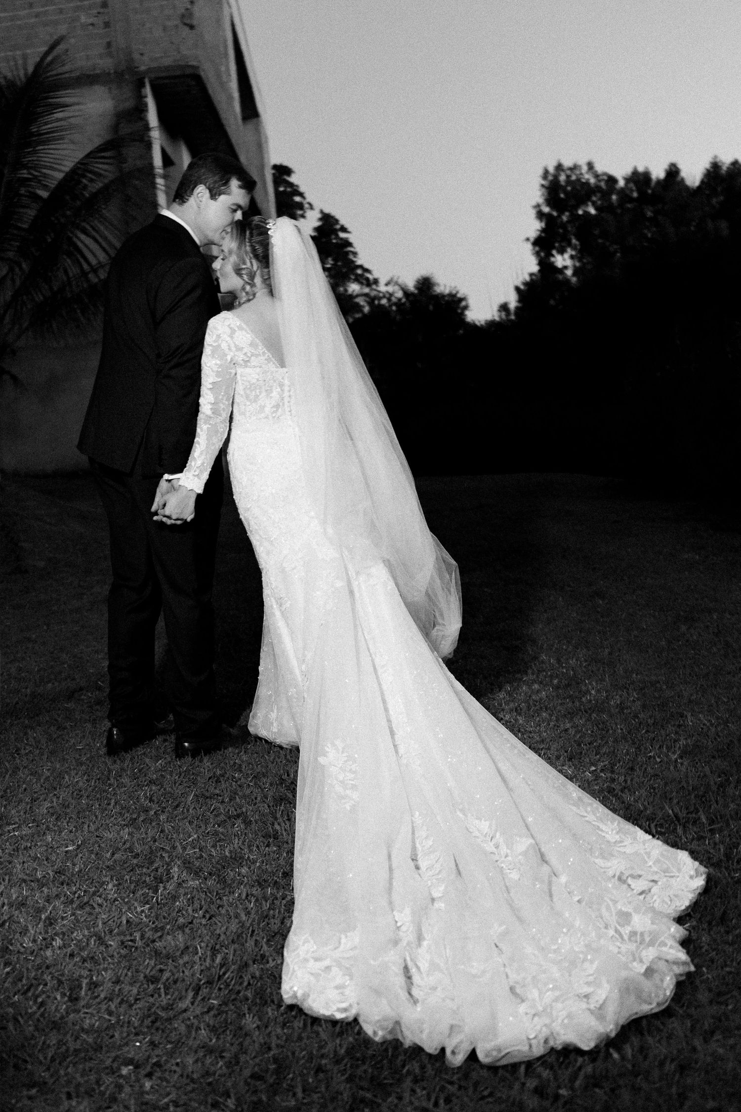 Black and white photo of bride in lace gown with long veil and groom in tuxedo sharing a romantic moment outdoors.