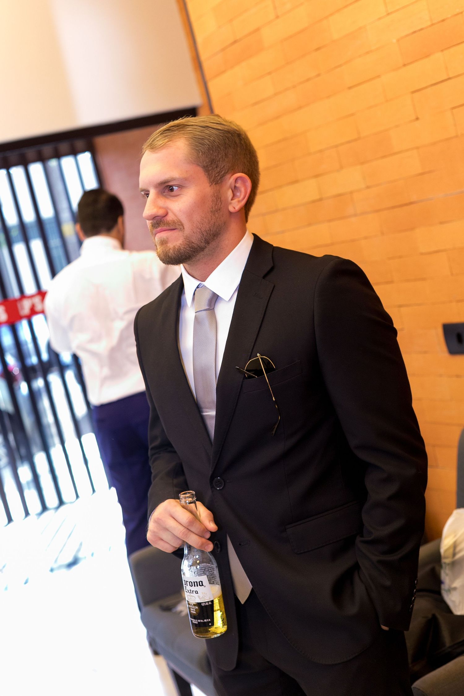 A man in a black suit holding a beer bottle at a formal event, with a brick wall background.
