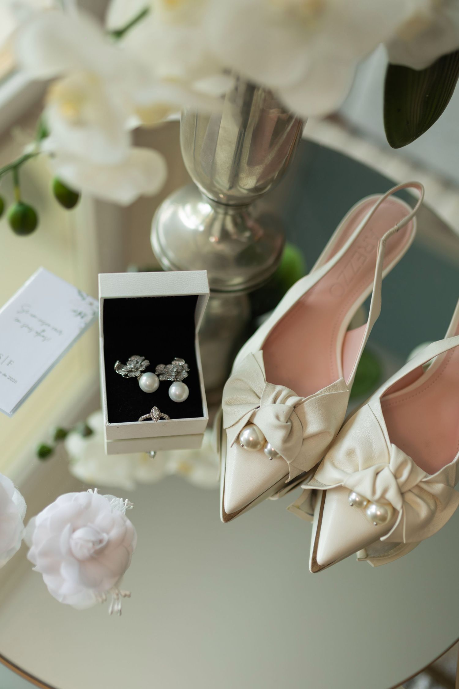 Elegant bridal accessories including pearl-embellished satin heels, diamond earrings, and a ring in a white jewelry box.
