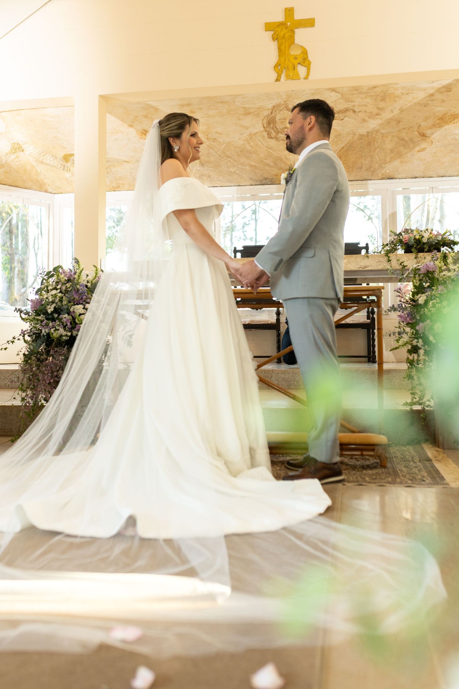Bride and groom hold hands at church altar during wedding ceremony, bride in white gown with veil, groom in gray suit.
