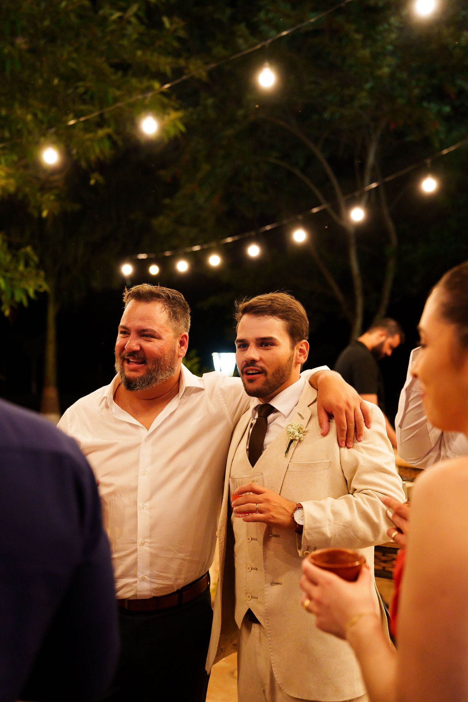 Groom in linen suit laughing with guest at outdoor wedding reception with string lights at night.