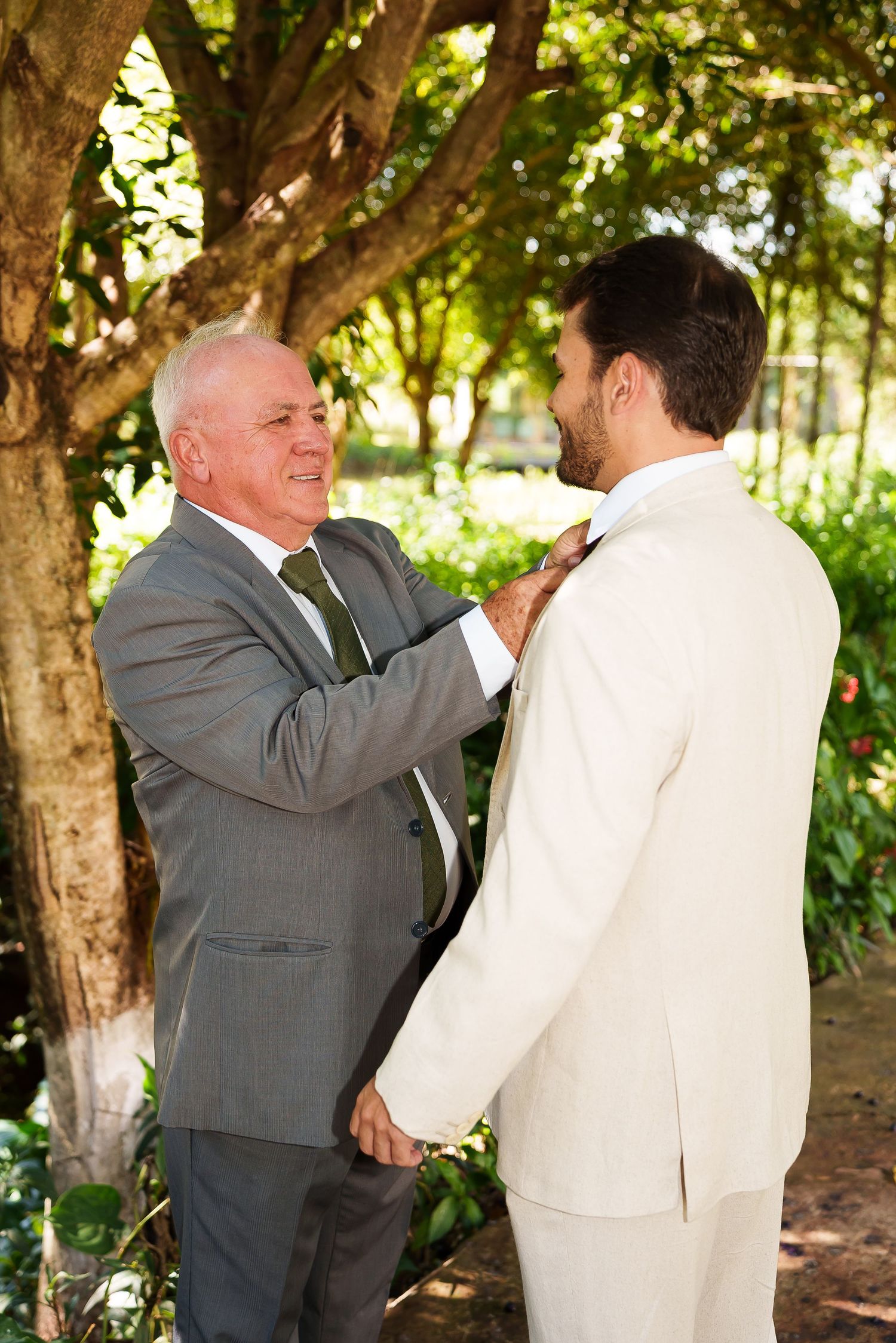 Older man in gray suit adjusting tie of young groom in cream suit outdoors before wedding ceremony.