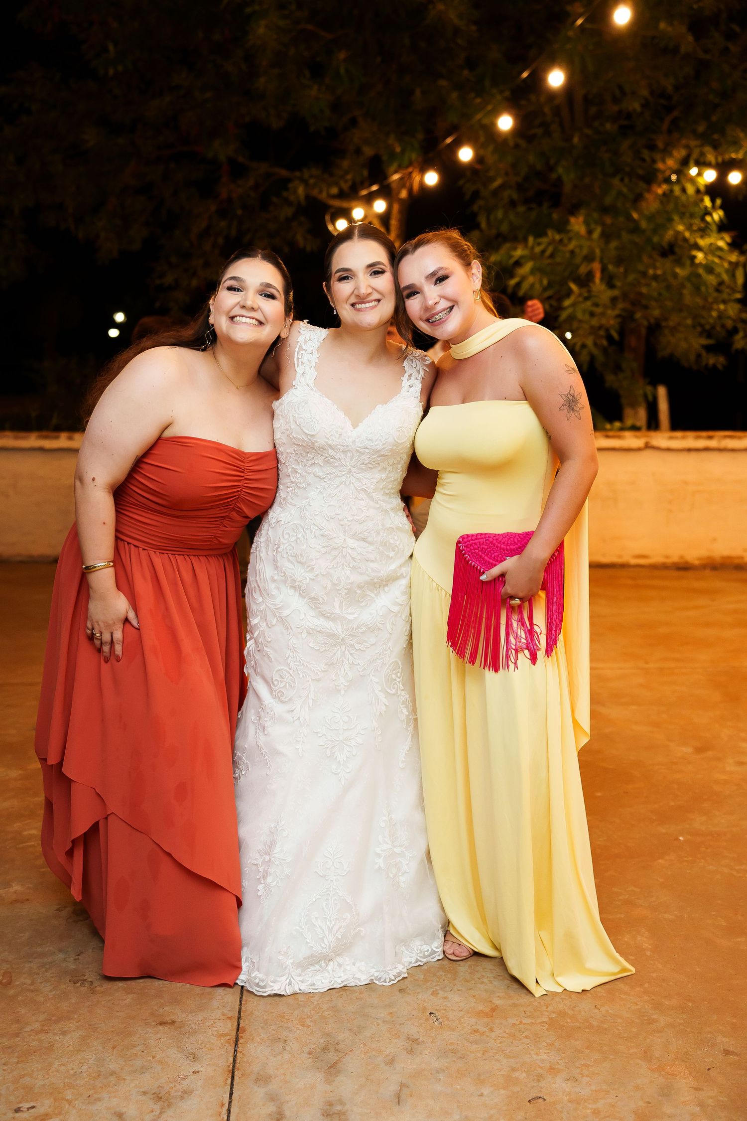Bride in white lace gown poses with two bridesmaids in red and yellow dresses at outdoor evening wedding reception.