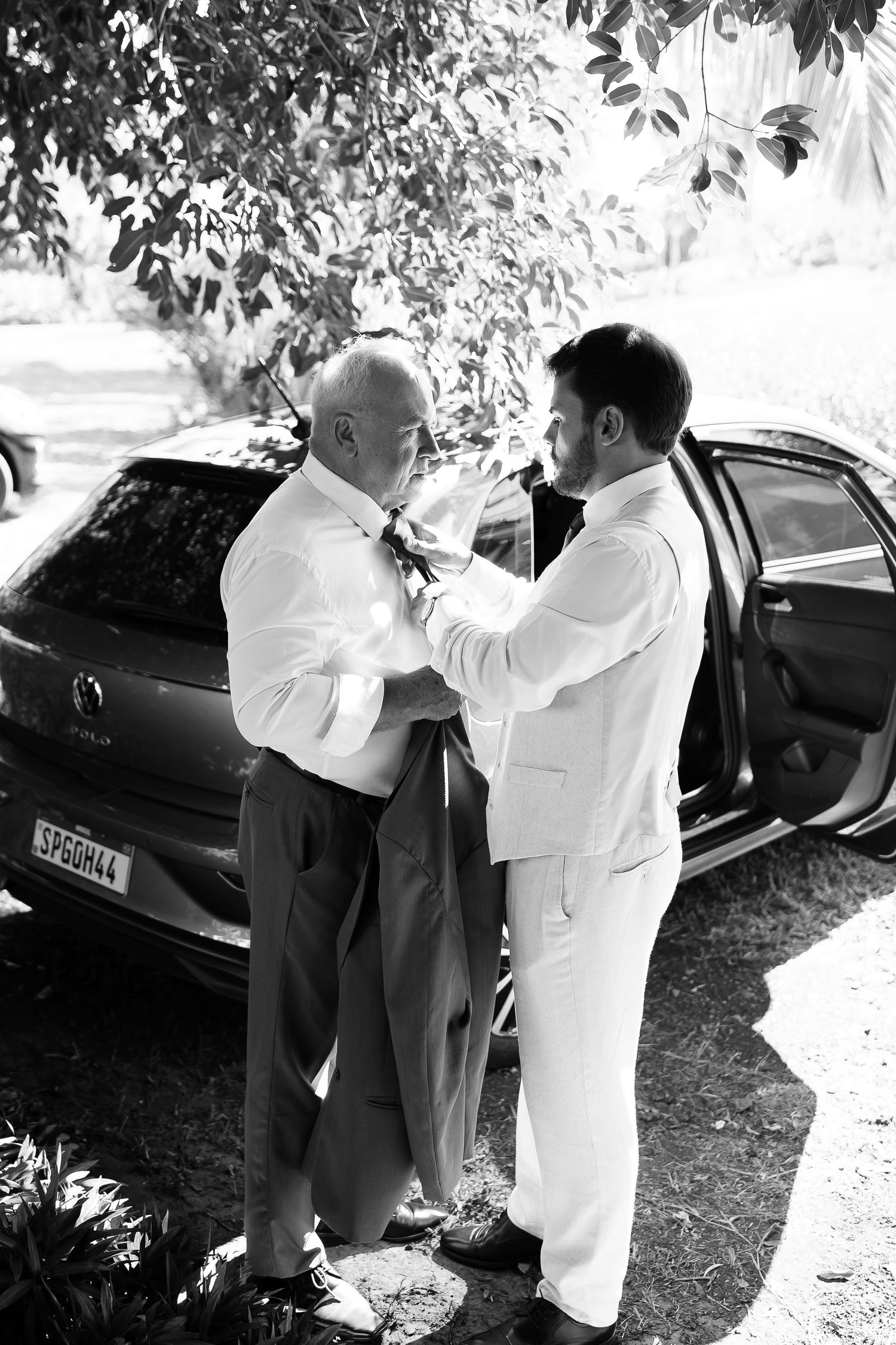 Black and white photo of groom in white suit helping older man adjust tie beside a car outdoors.