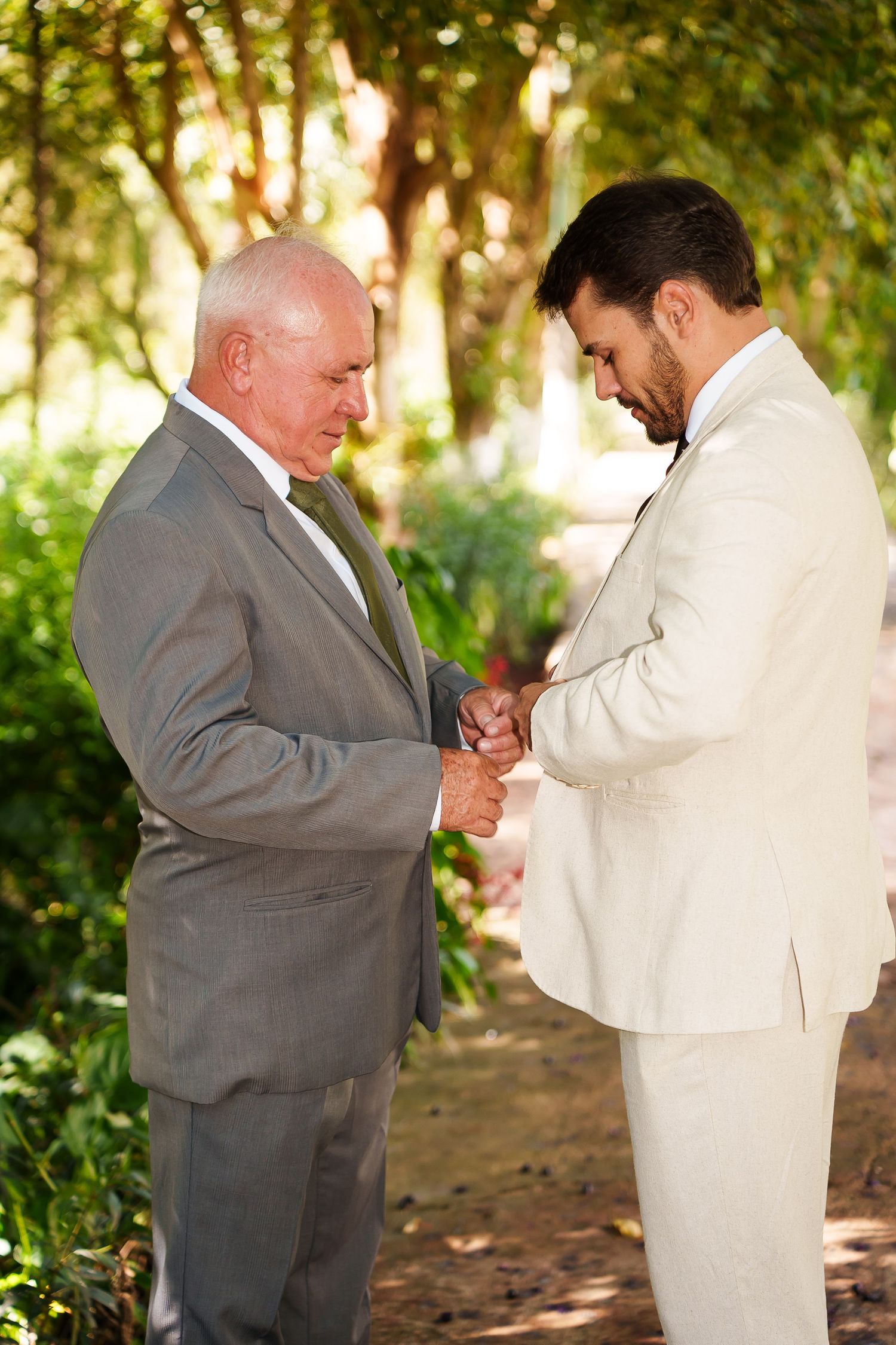 Two men in suits shaking hands outdoors at a wedding ceremony surrounded by lush green trees.
