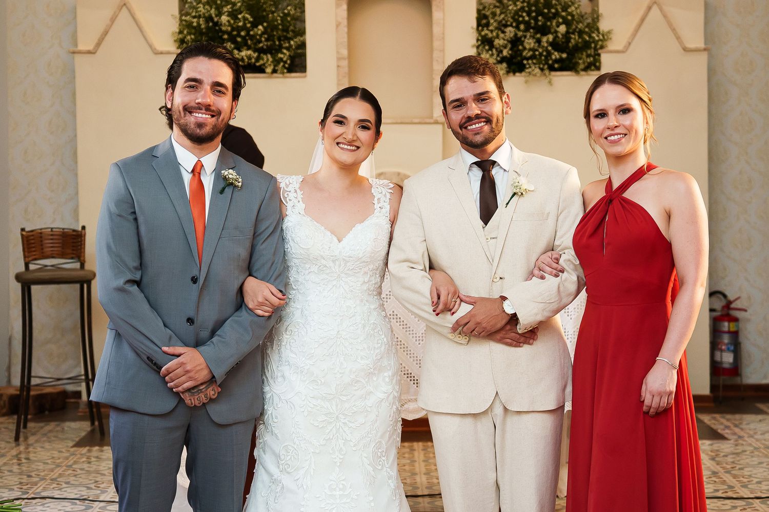 Wedding party posing outdoors, featuring bride in white lace gown, groom in cream suit, groomsman, and bridesmaid in red dress.