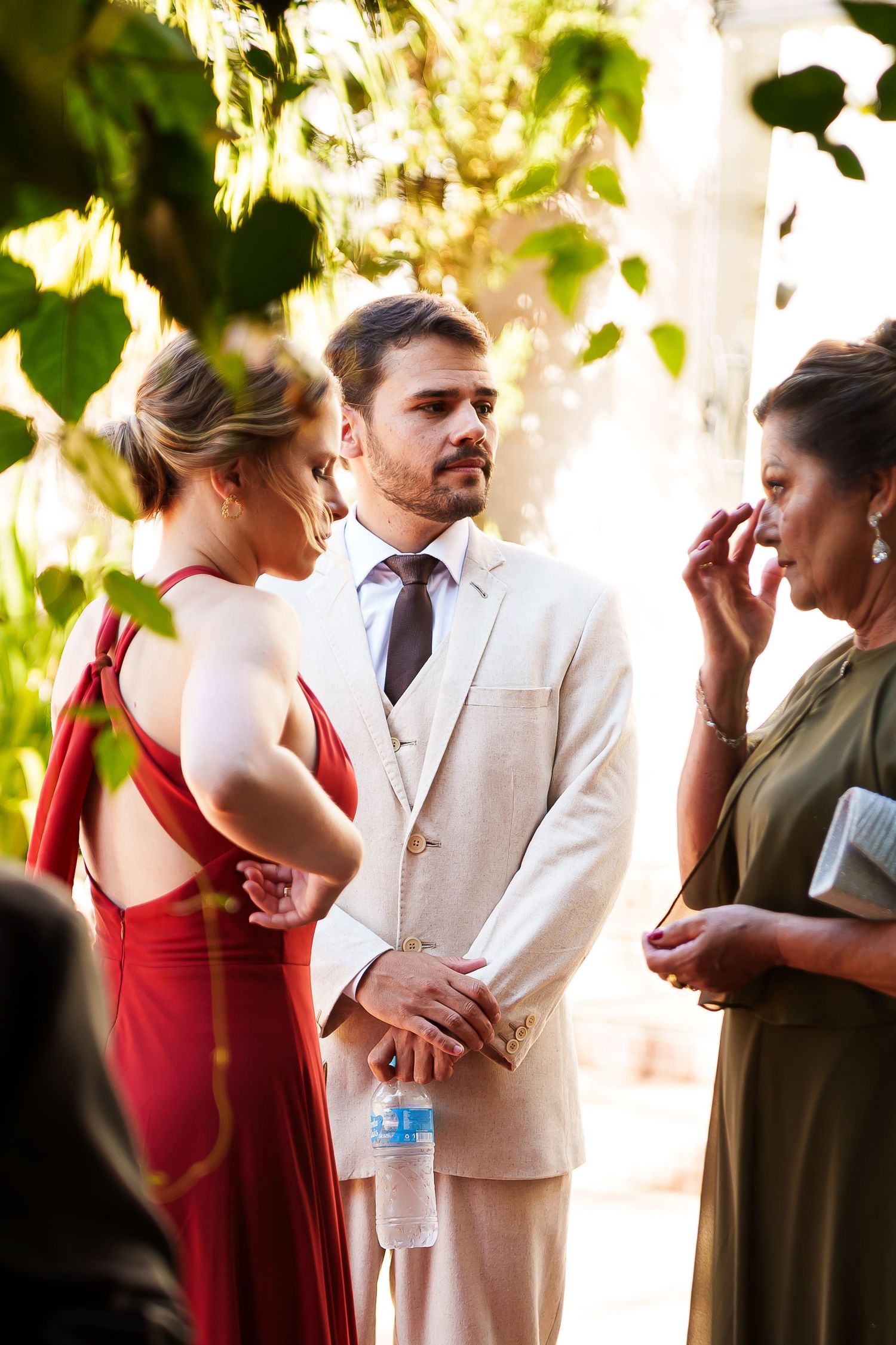 Groom in white suit stands with bridesmaids at outdoor wedding ceremony, emotional guest wiping tears nearby.