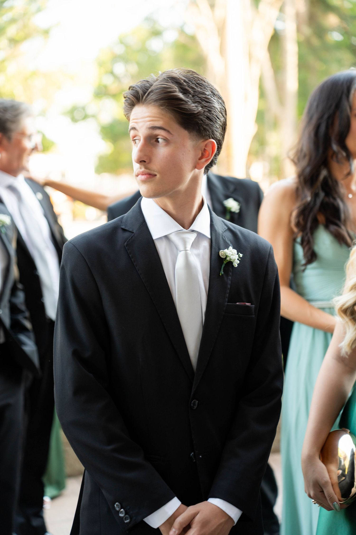 Young man in black tuxedo with white tie and boutonniere at a wedding ceremony outdoors.
