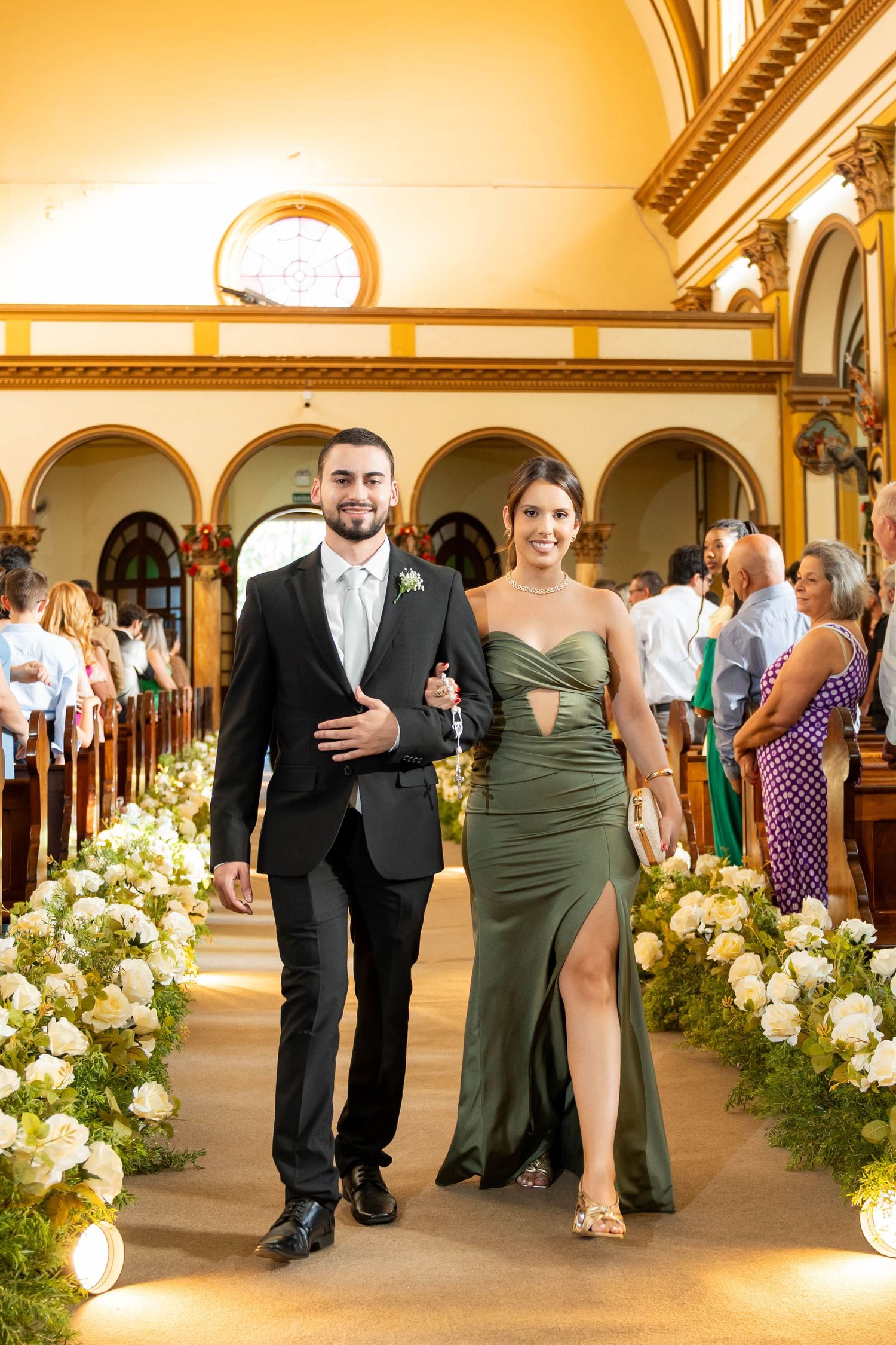Groomsman in black tuxedo escorts bridesmaid in elegant green gown down floral-lined church aisle.