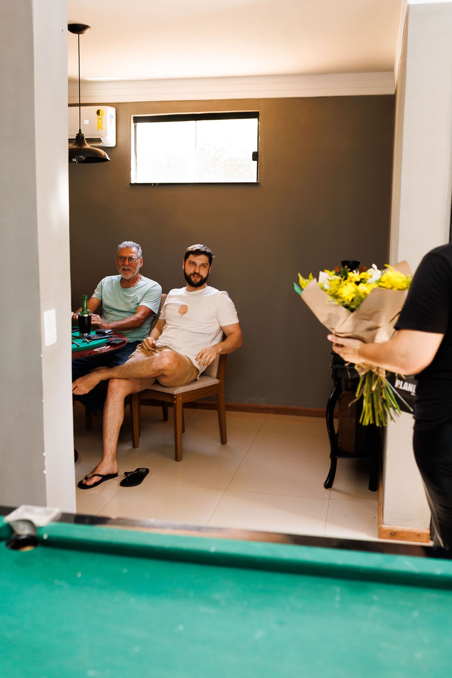 Two men relaxing in a game room near a pool table while a person holds a bouquet of yellow flowers.