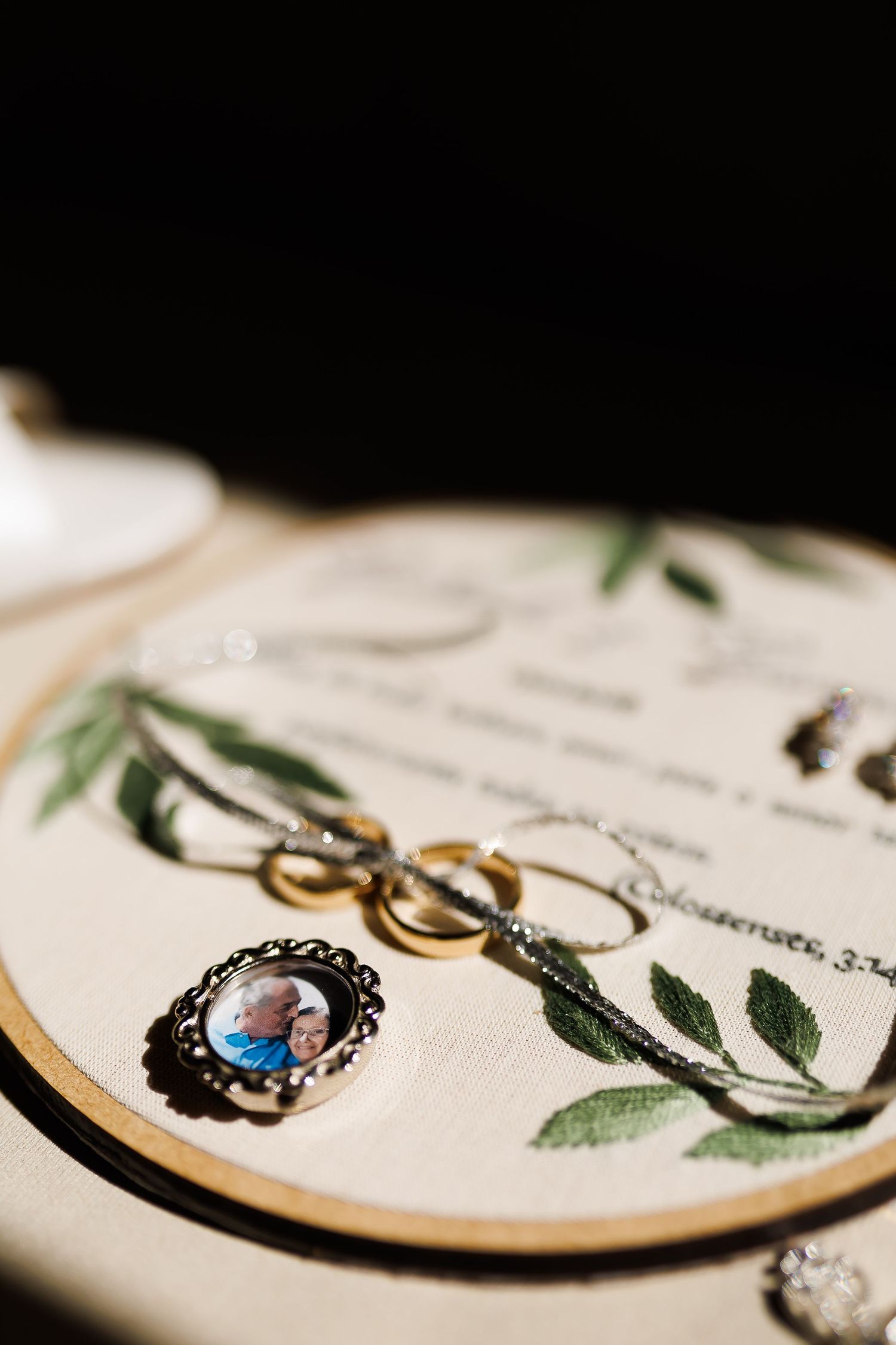 Wedding rings, a photo brooch, and a necklace arranged on an embroidered hoop with greenery and a Bible verse.