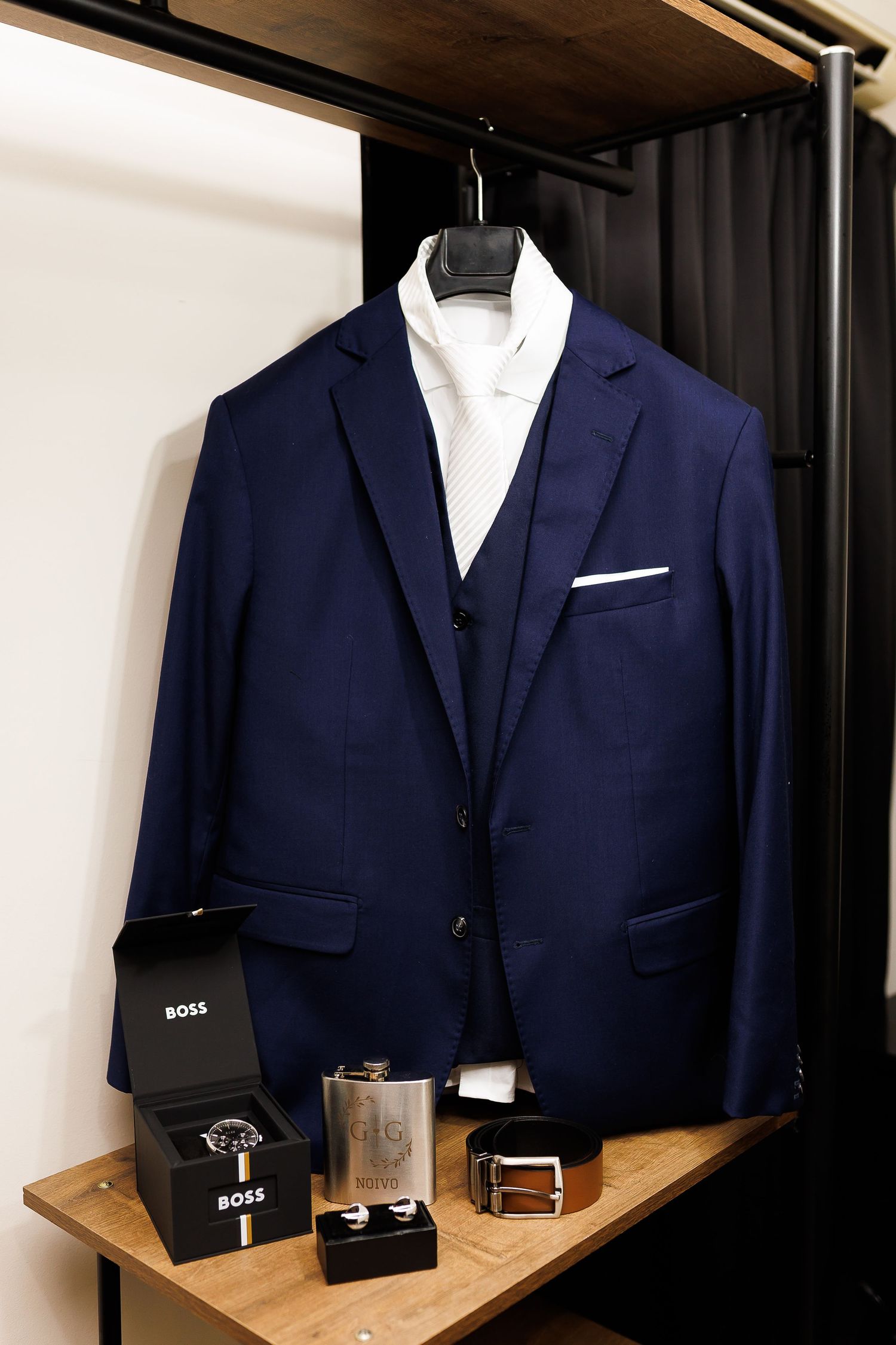 Navy blue Hugo Boss suit jacket displayed on hanger with white shirt, watch, flask, belt, and cufflinks on wooden shelf.