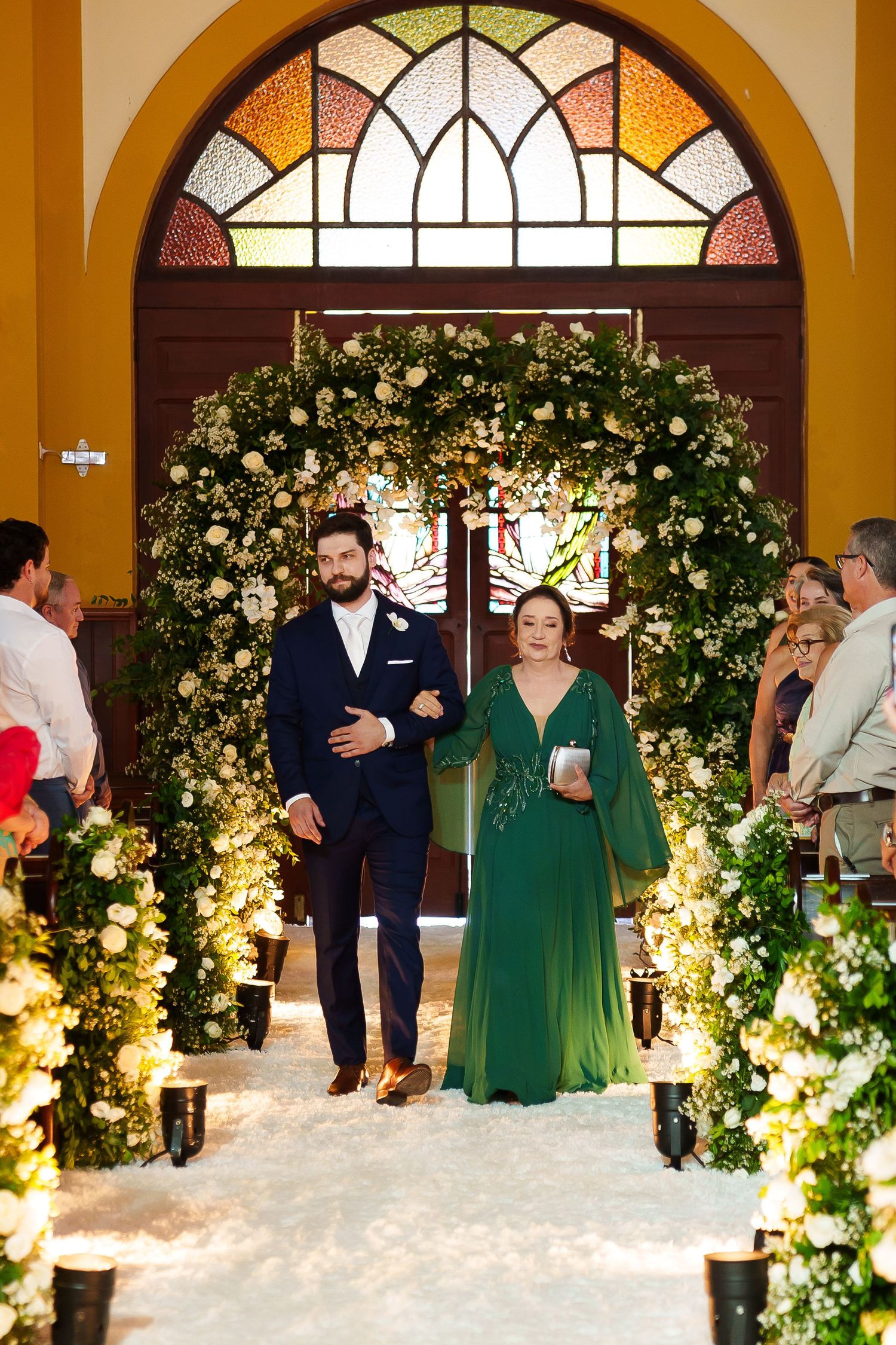 Groom in navy suit escorts woman in emerald green gown down floral-adorned wedding aisle with white flower arch.