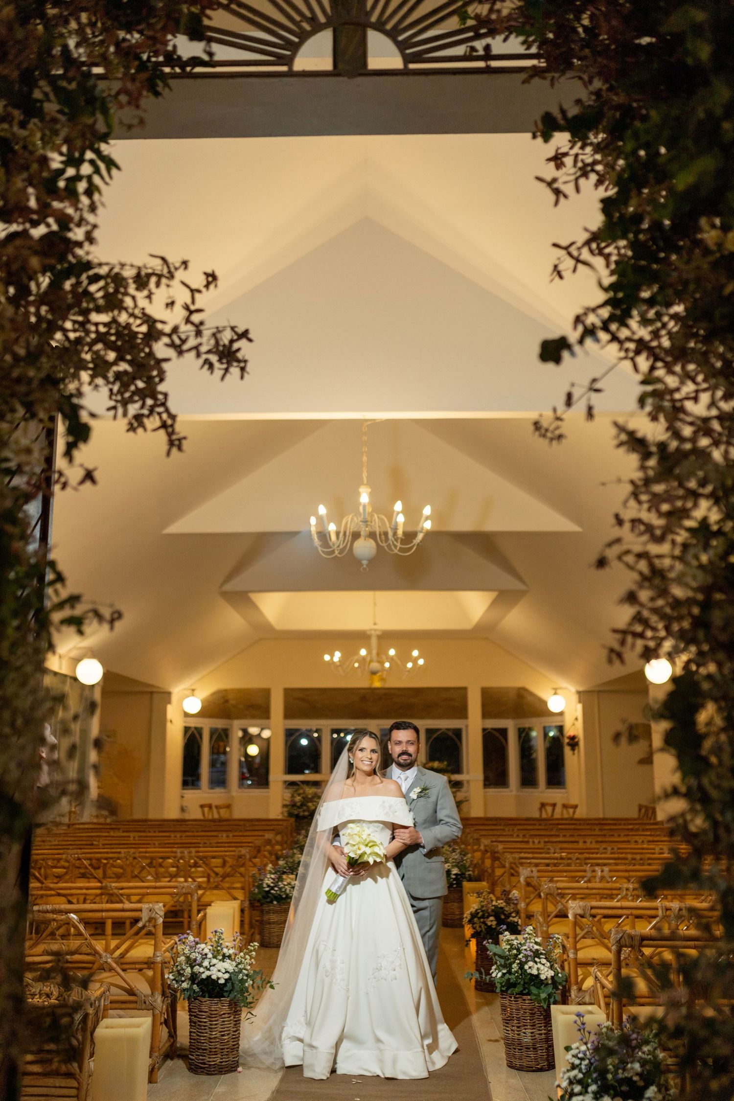 Bride and groom stand together in an elegantly decorated chapel with wooden pews, floral arrangements, and warm chandelier lighting.