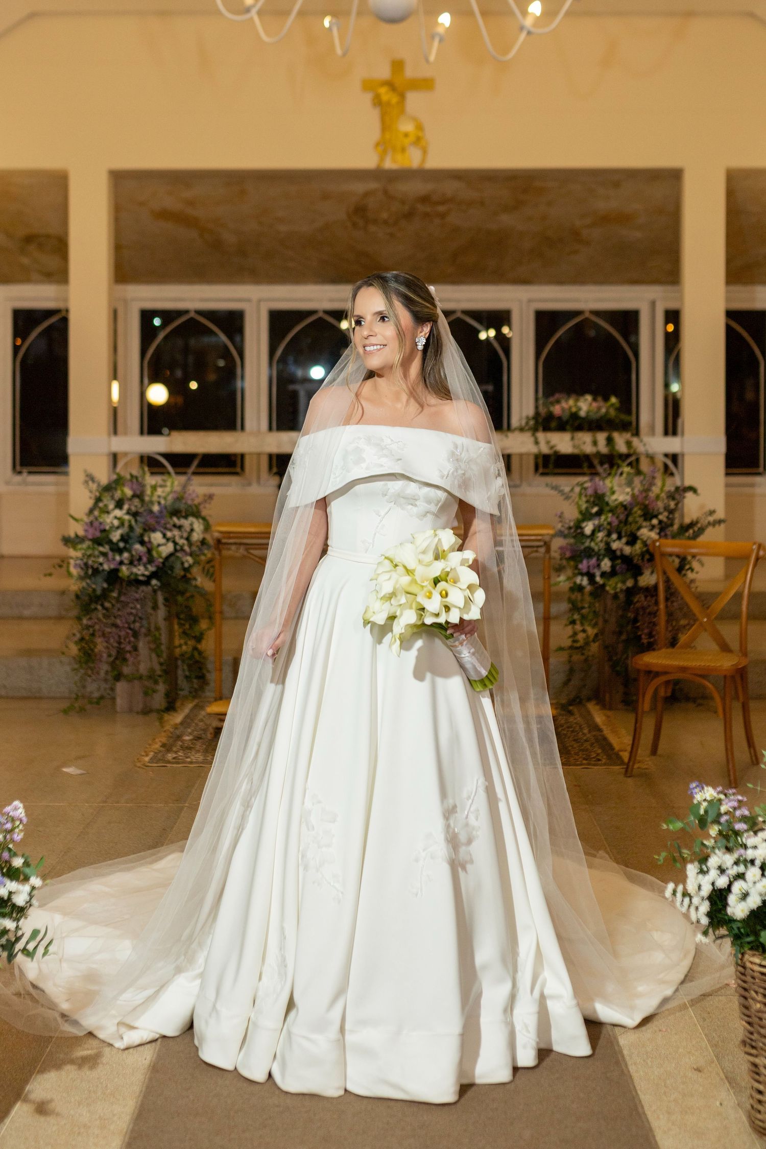Bride in off-shoulder white ball gown with veil holds bouquet inside decorated church aisle.