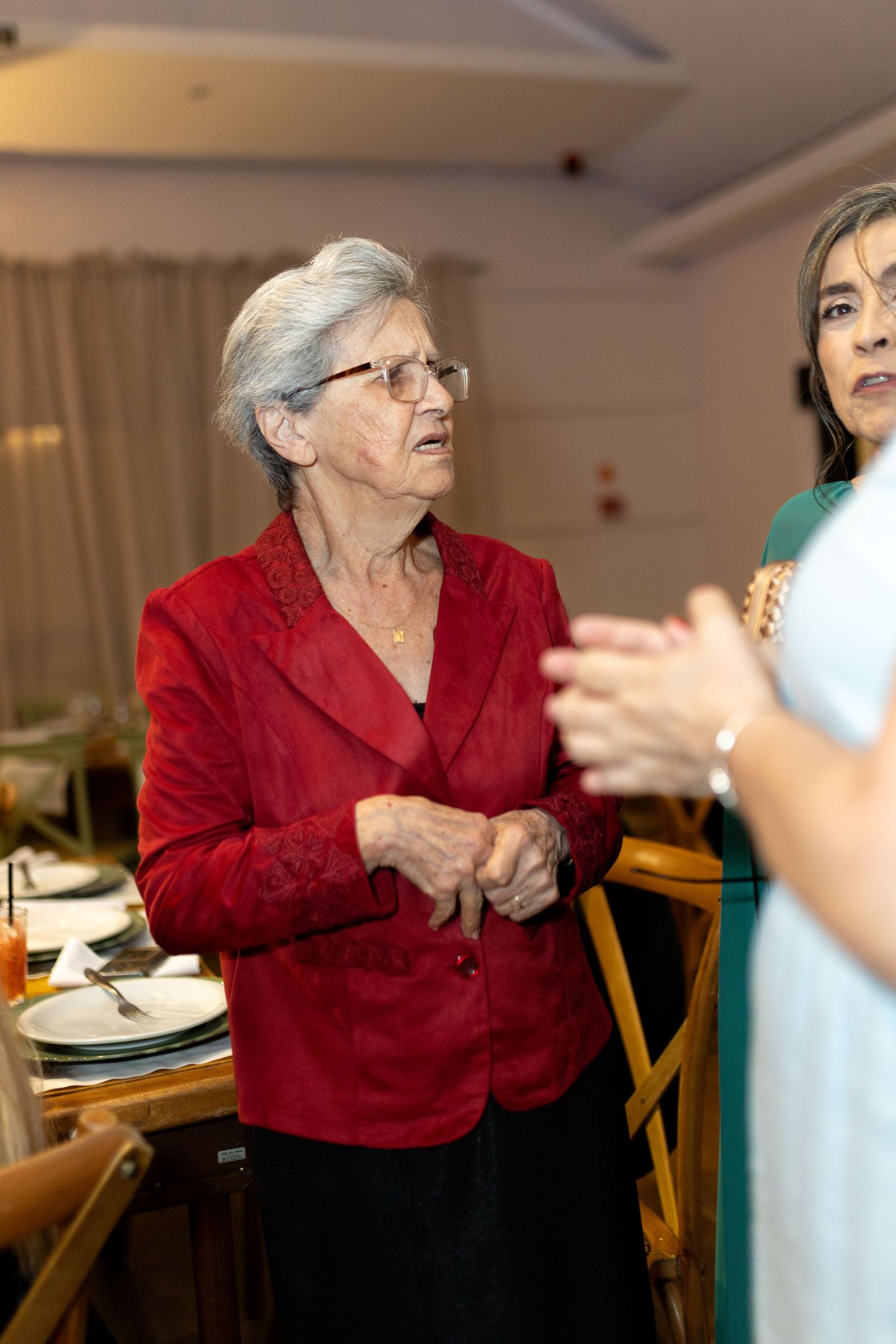 Elderly woman in red jacket conversing at a formal dinner event with another guest nearby.
