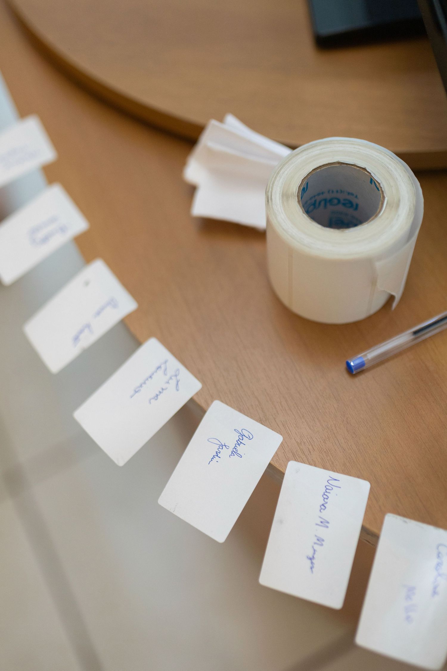 Name tags with handwritten labels arranged on a wooden table beside a roll of label tape and a blue pen.