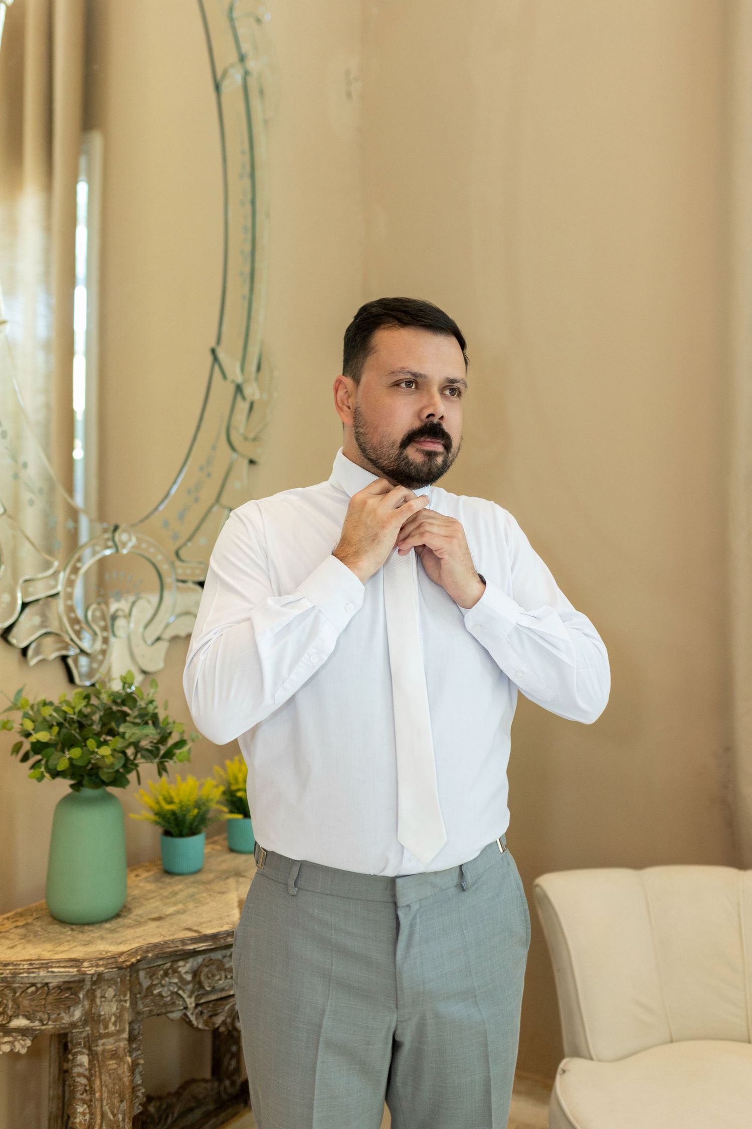 Bearded groom in white shirt tying his necktie in a warmly lit room with mirror.