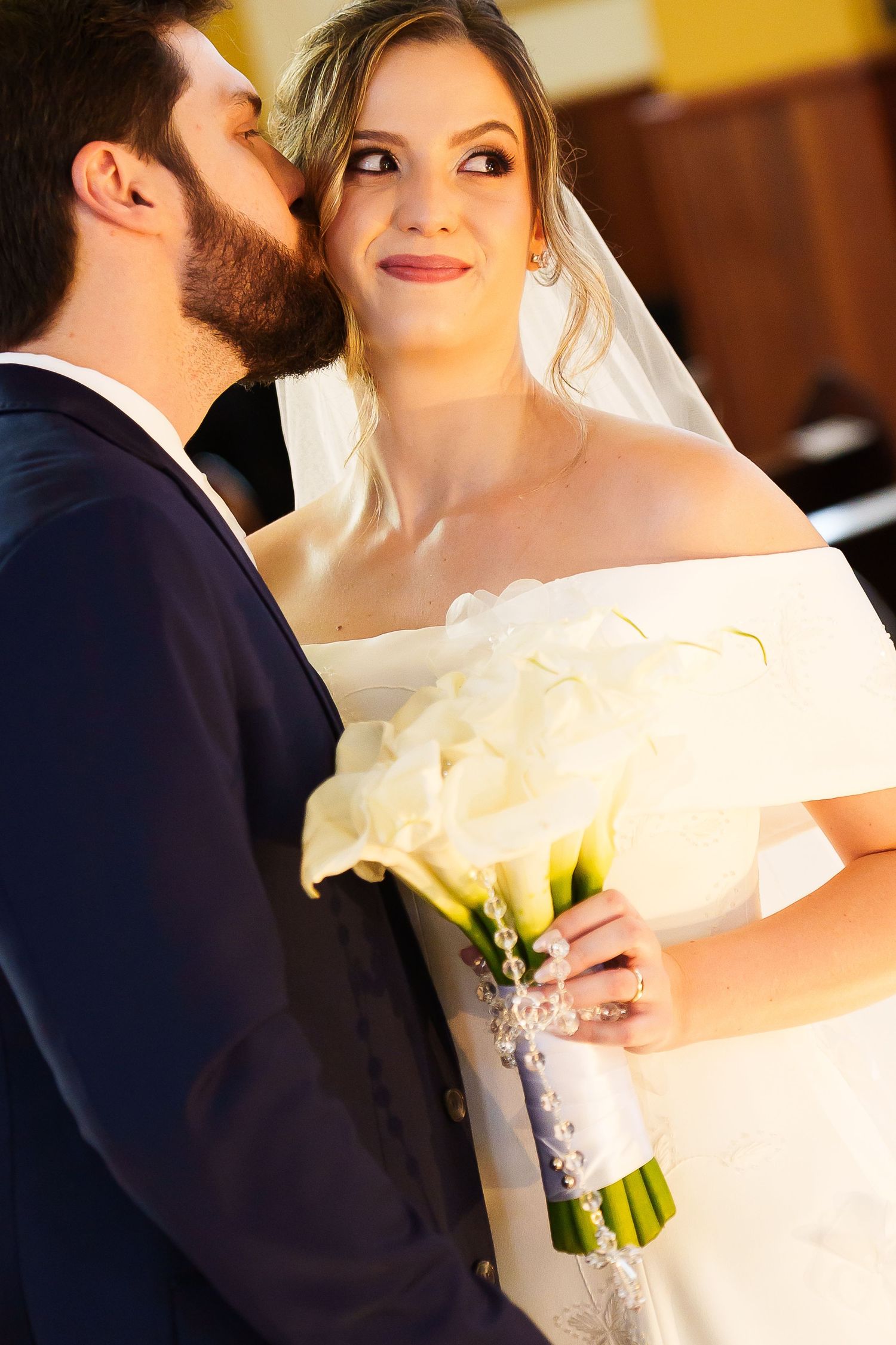 Groom in navy suit kissing bride in white off-shoulder gown holding calla lily bouquet on their wedding day.