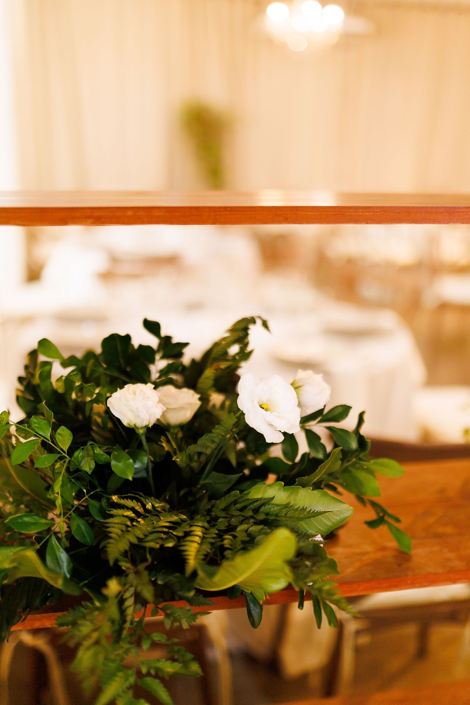 Elegant white floral arrangement with lush green foliage on wooden railing in a softly lit wedding reception venue.