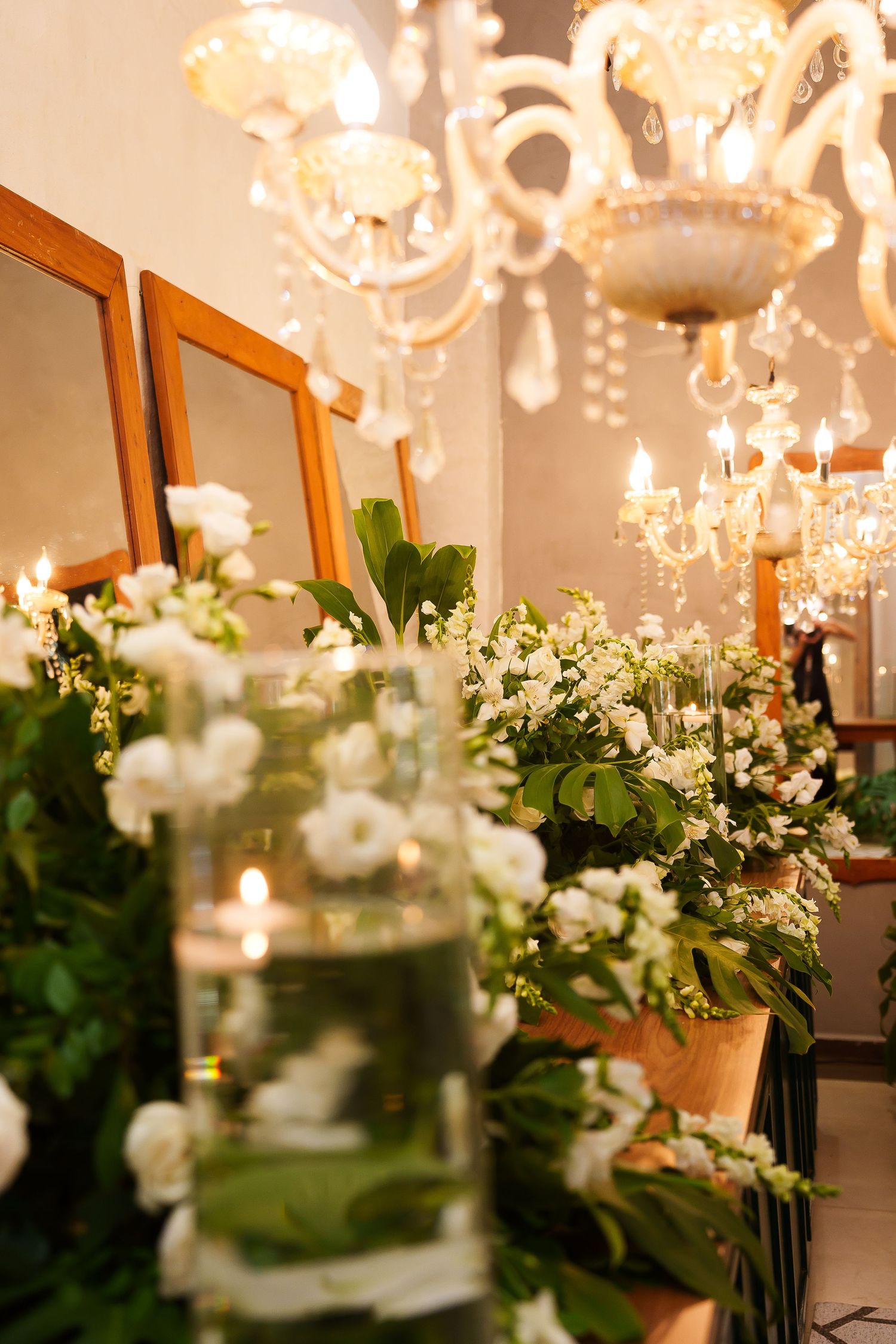 Elegant floral decor with white roses, greenery, candles in glass vases, and crystal chandeliers in a warmly lit room.