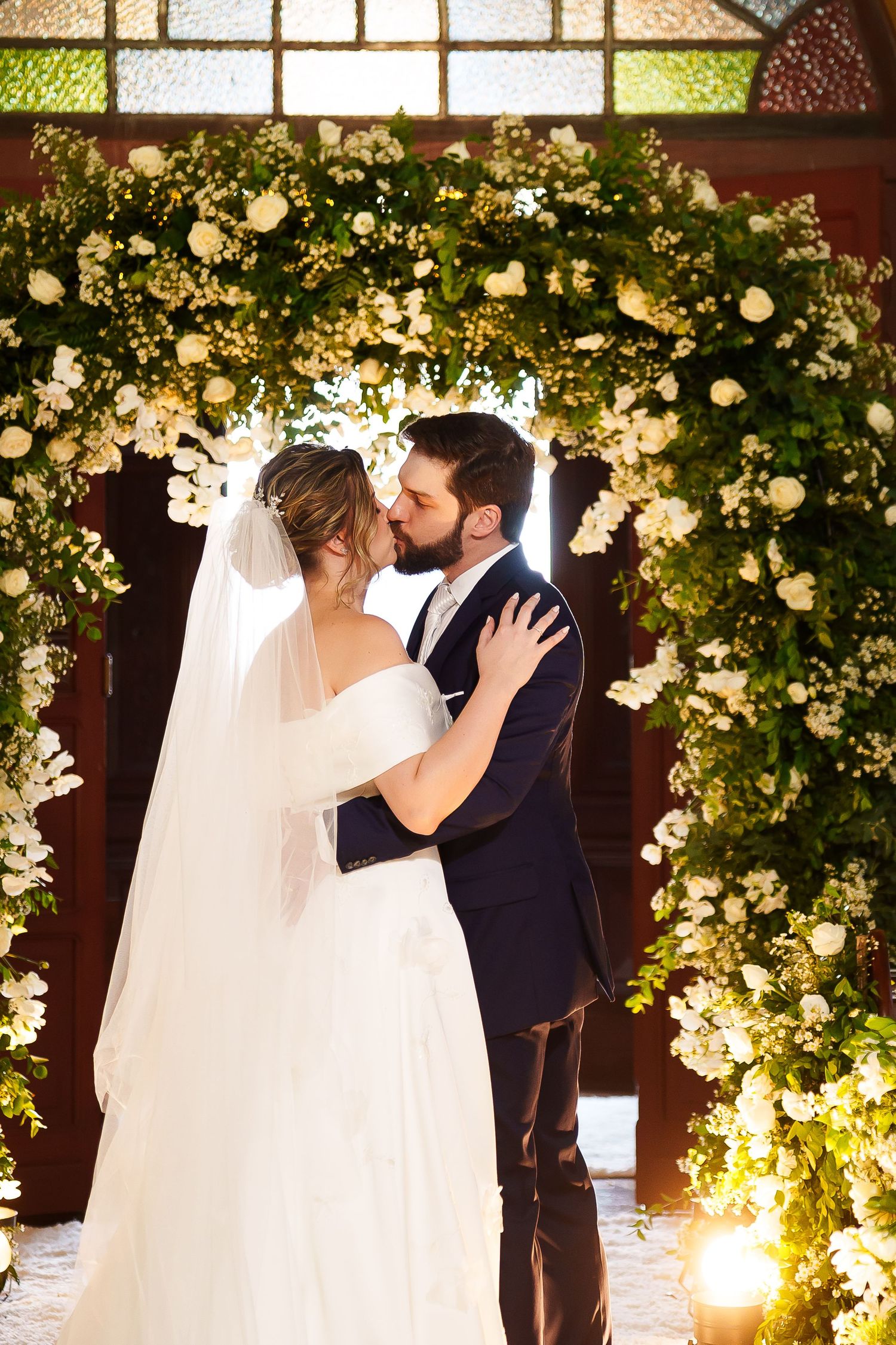 Bride and groom share first kiss under floral arch with white roses and greenery during wedding ceremony.