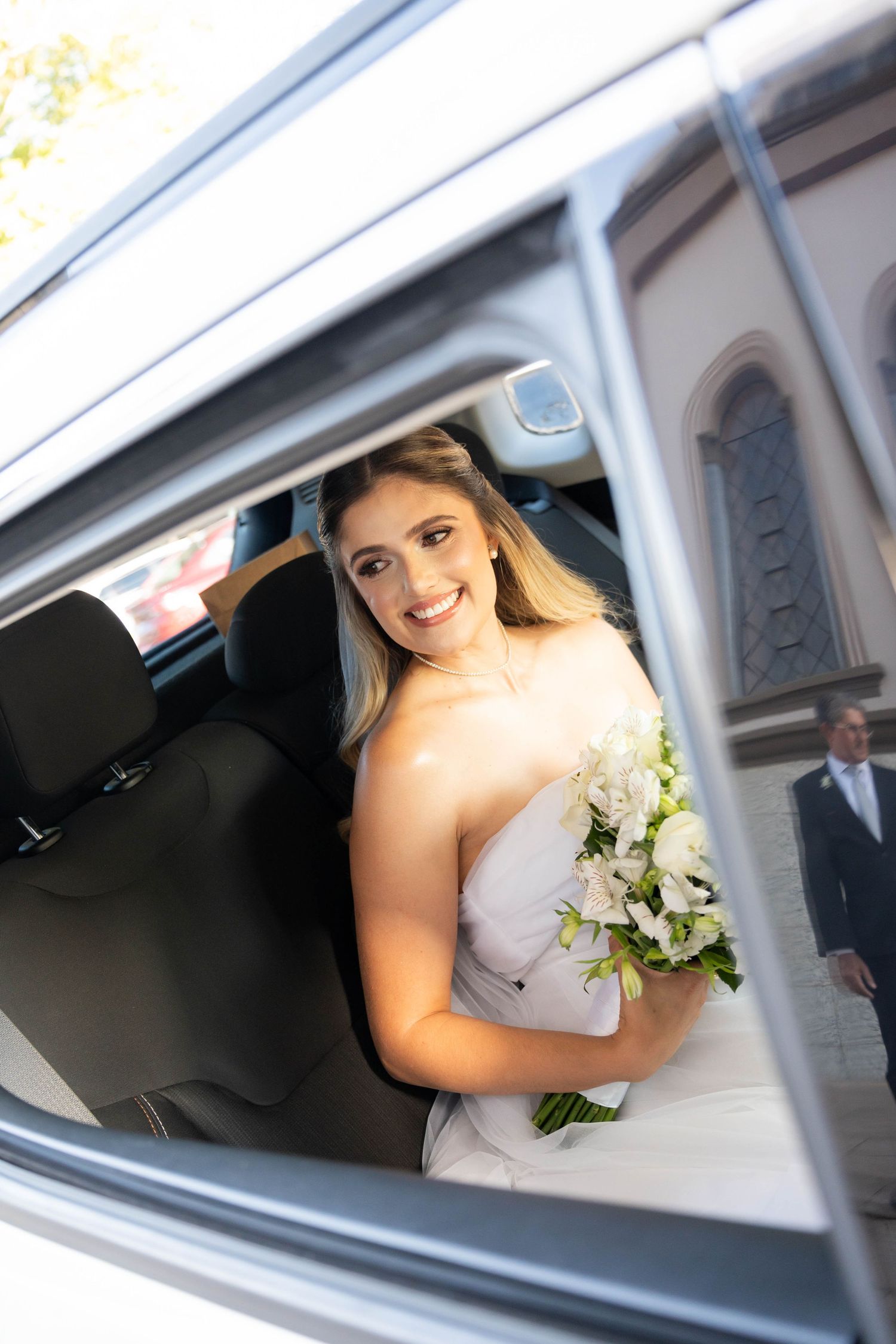 Smiling bride in white strapless gown holding white flower bouquet stepping out of a luxury car on her wedding day.