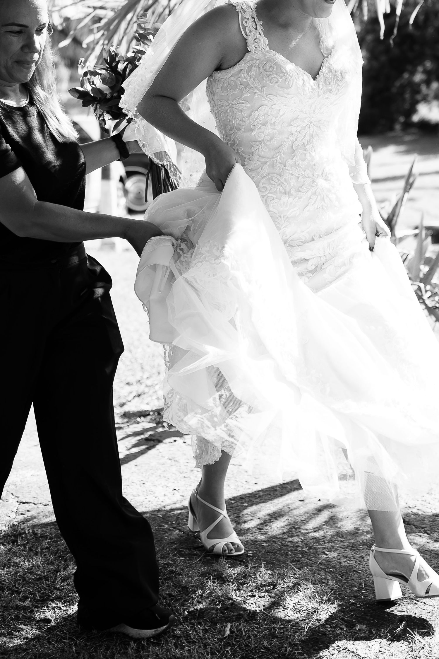 Black and white photo of a bride in lace gown with helper adjusting dress train outdoors.