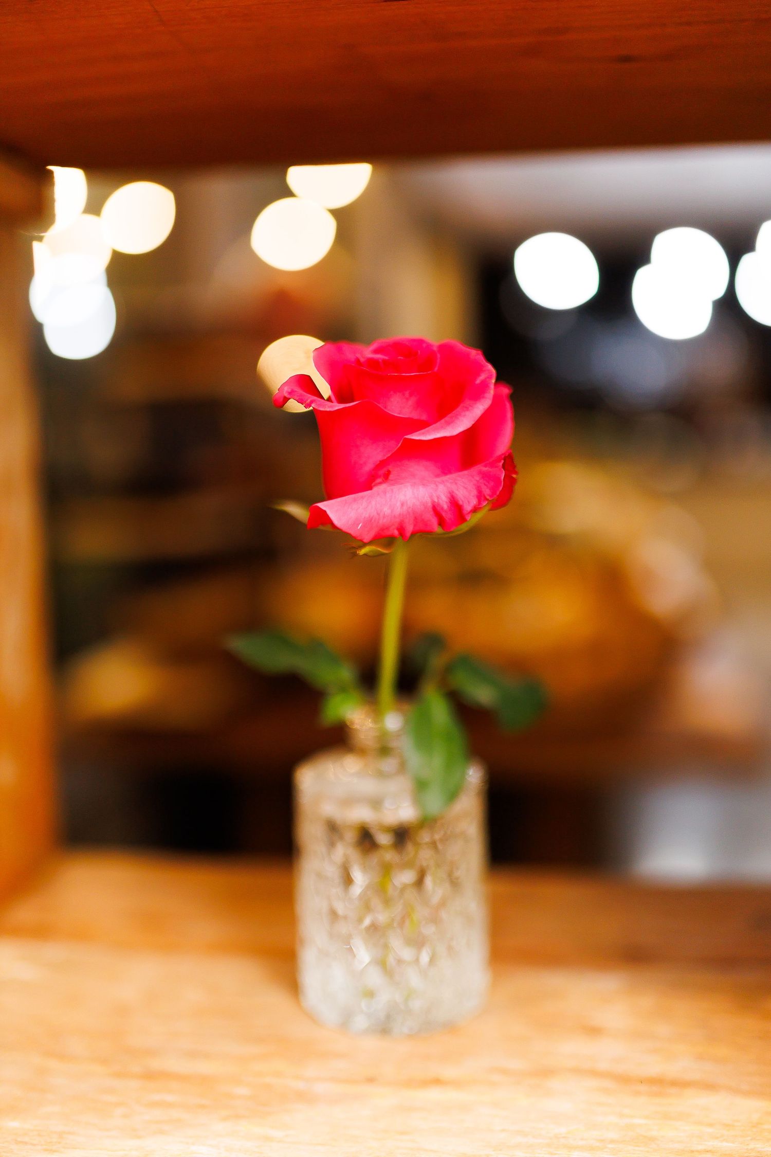 A single red rose in a small glass vase on a wooden surface with bokeh lights in the background.