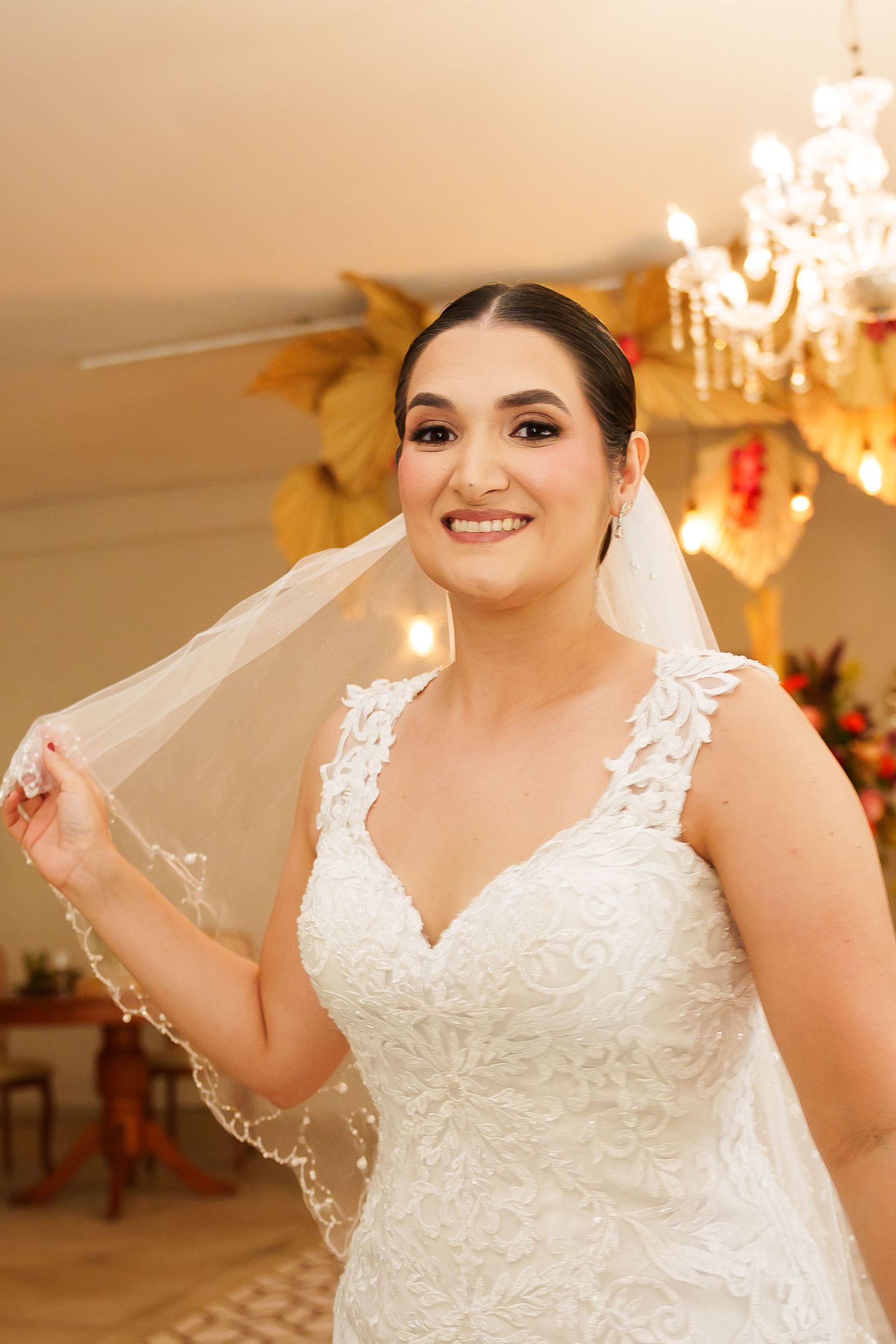 Smiling bride in lace wedding dress holding veil, standing under chandelier in warmly lit venue.