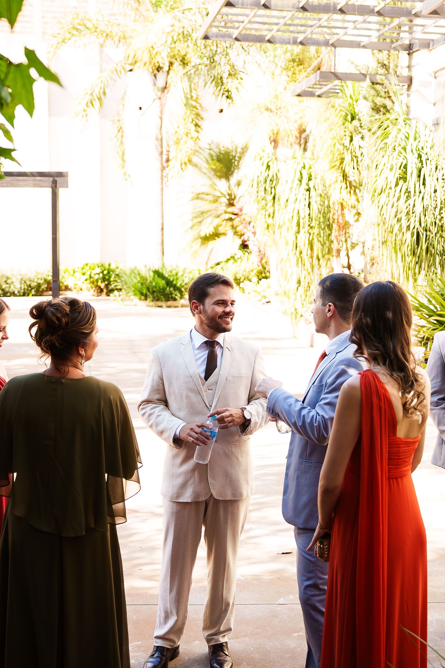 Elegantly dressed wedding guests socializing outdoors, with a smiling groom in a beige suit at a sunny garden venue.