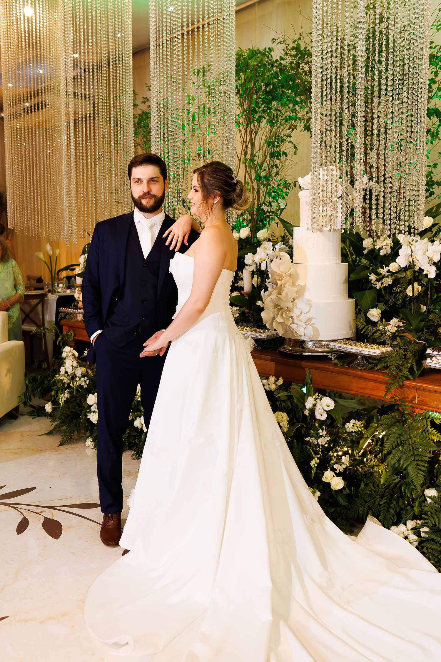 Elegant bride and groom posing near a white wedding cake surrounded by lush greenery and crystal decor.