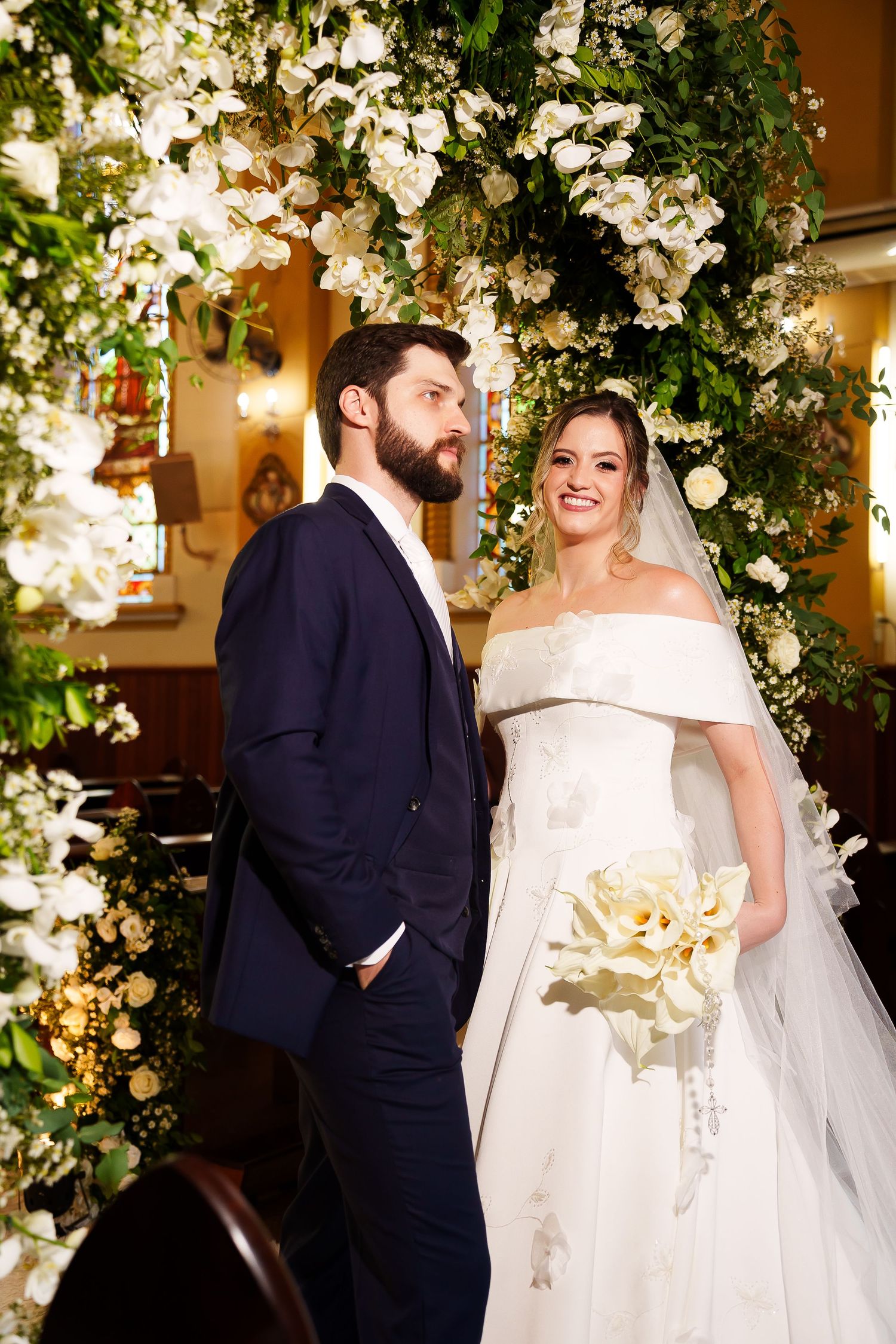 Bride and groom stand under floral arch in church, bride holds white bouquet in elegant off-shoulder gown.