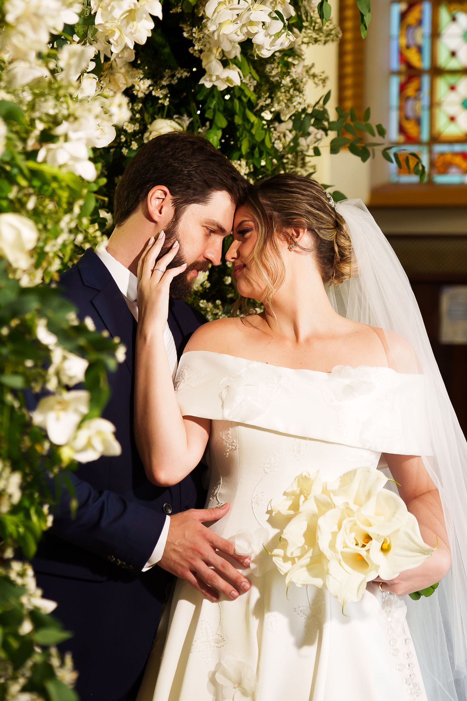 Bride and groom share a romantic moment at altar surrounded by white floral arrangements in a sunlit church.