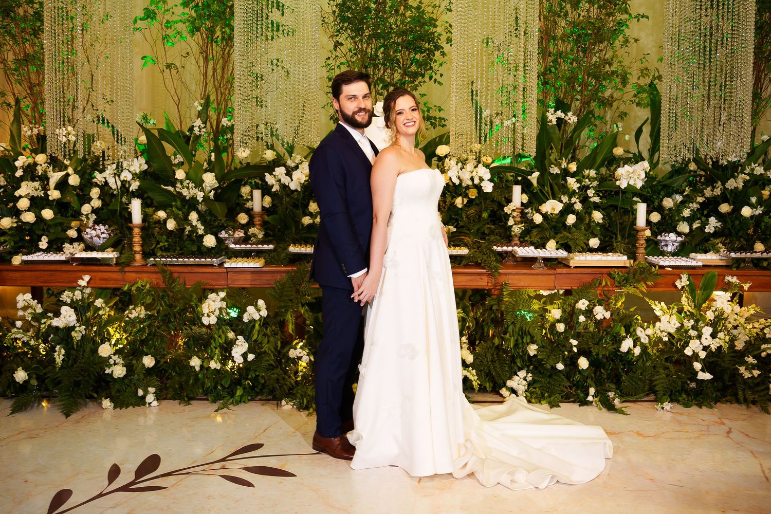 Bride and groom pose elegantly in front of a lush floral backdrop with white blooms at their wedding reception.
