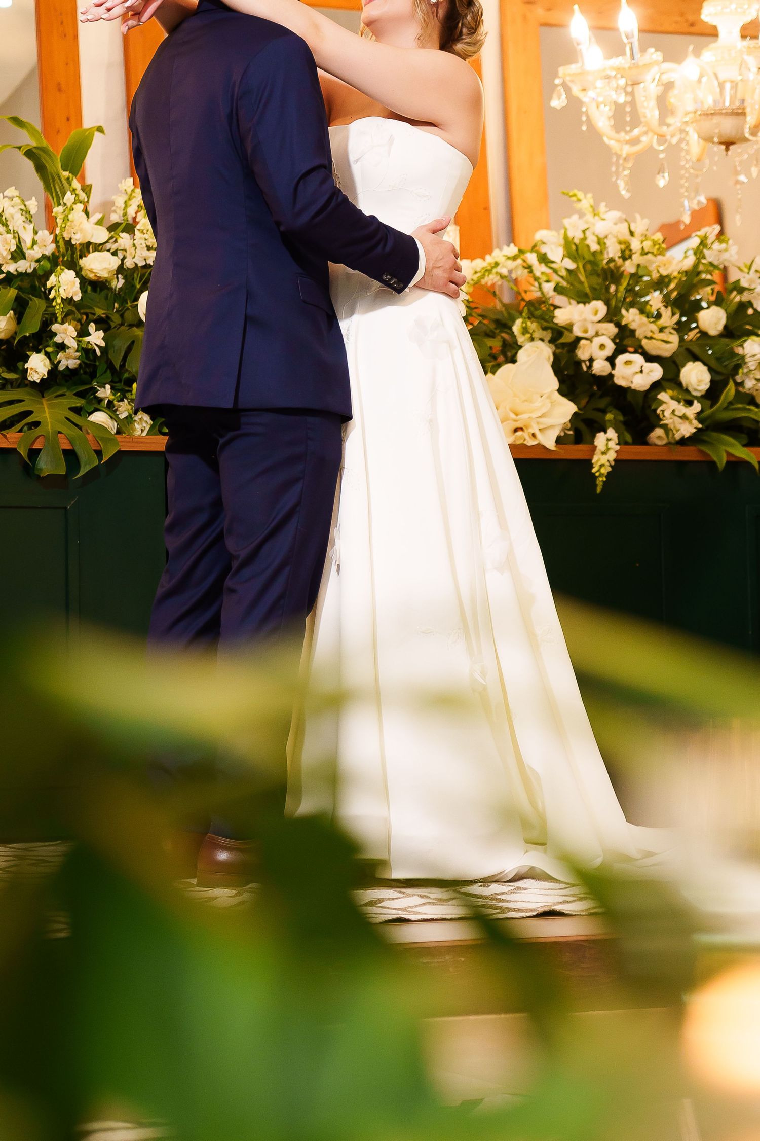 Bride in white gown and groom in navy suit embrace during wedding ceremony with floral arrangements in background.