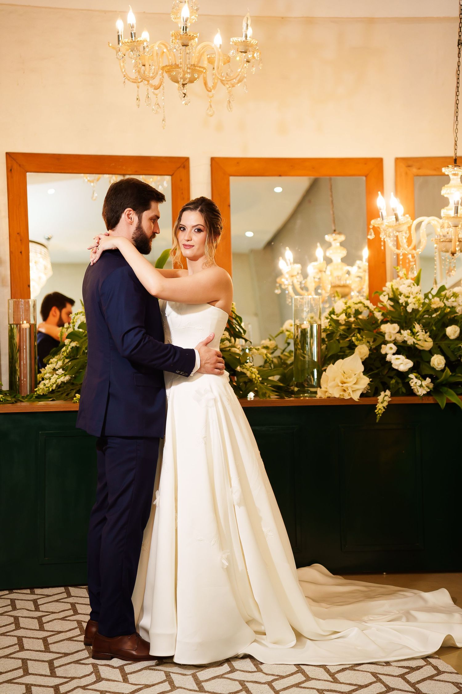 Elegant bride and groom embracing in a candlelit wedding venue with floral arrangements and gold chandelier.