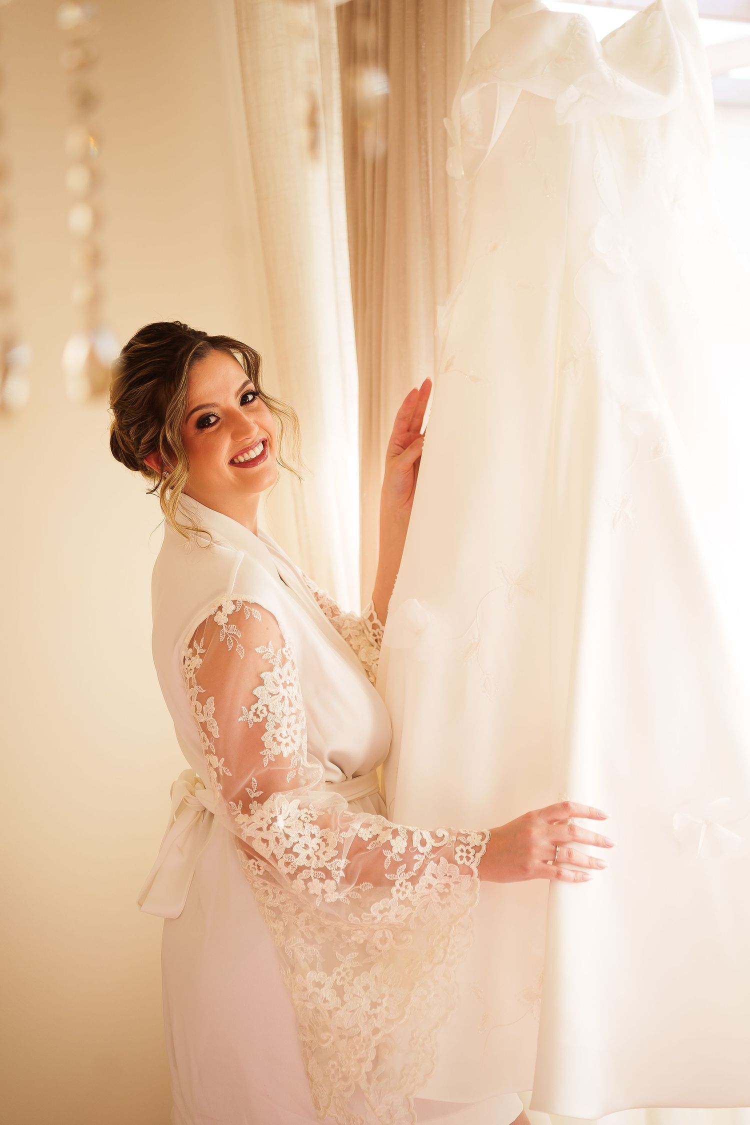 Smiling bride in white lace robe reaching for her wedding dress hanging by a window on her wedding day.