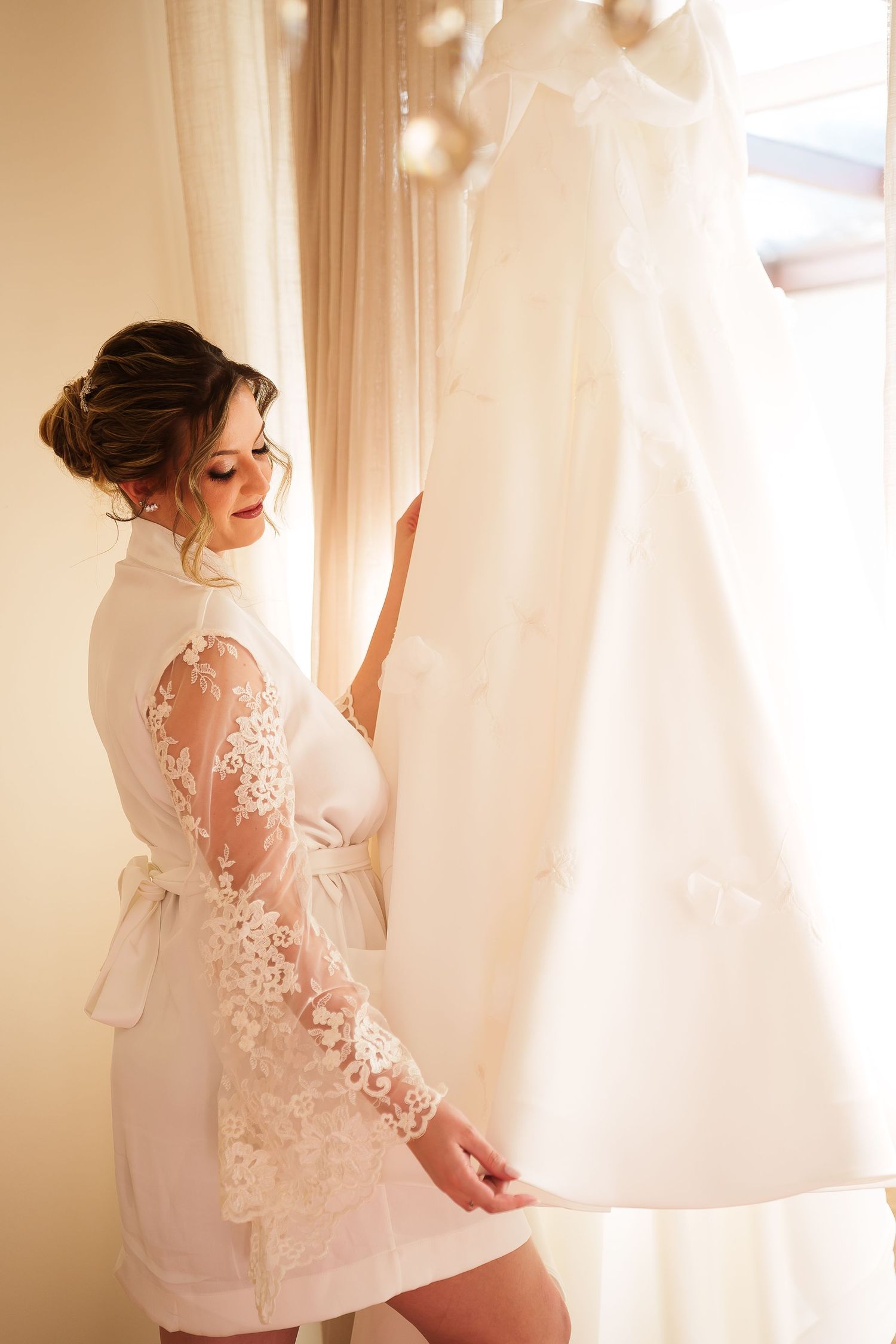 Bride in white lace robe admiring her wedding dress hanging by a window on her wedding morning.