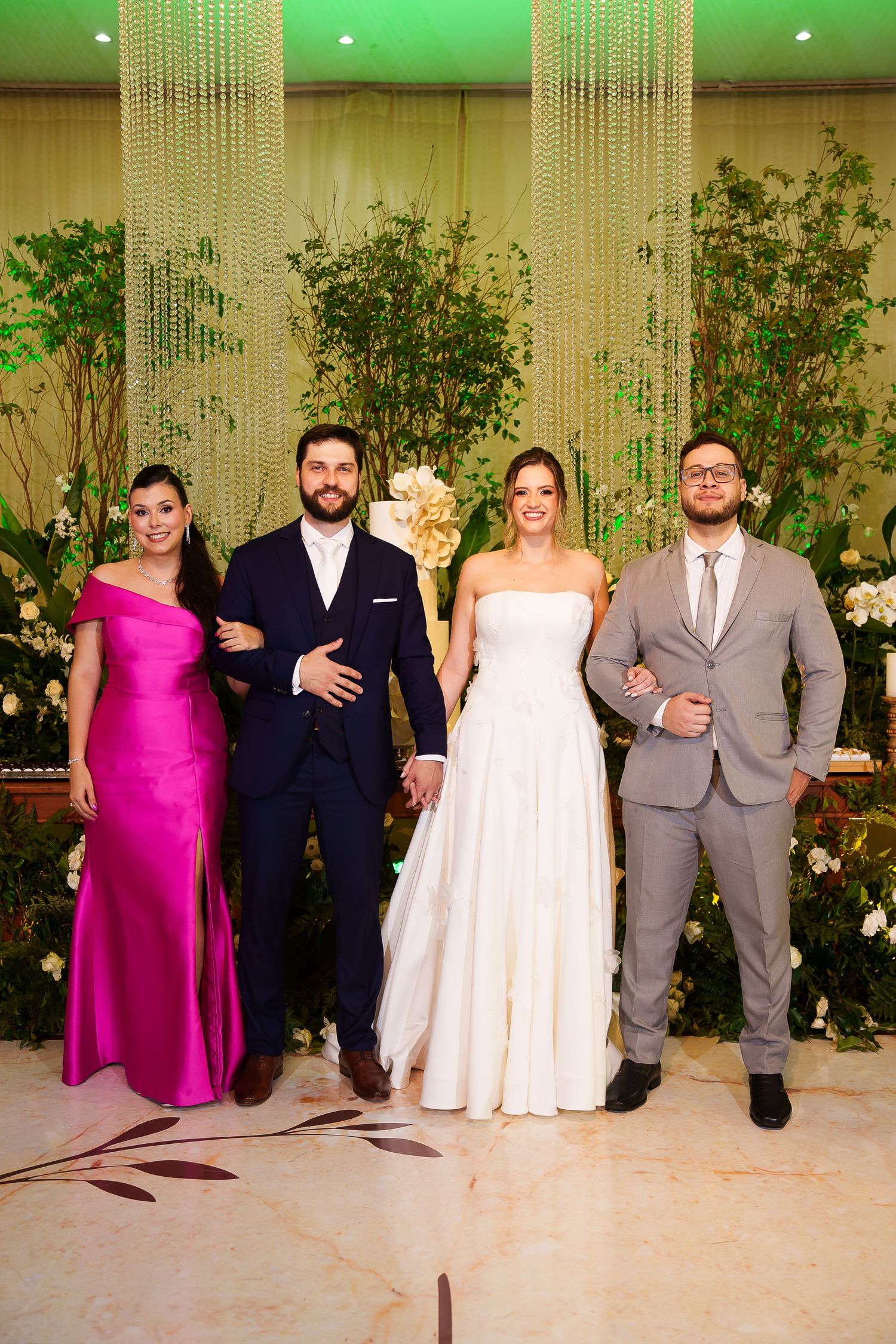 Wedding party of four posing elegantly, bride in white gown, groom in navy suit, guests in pink dress and gray suit.