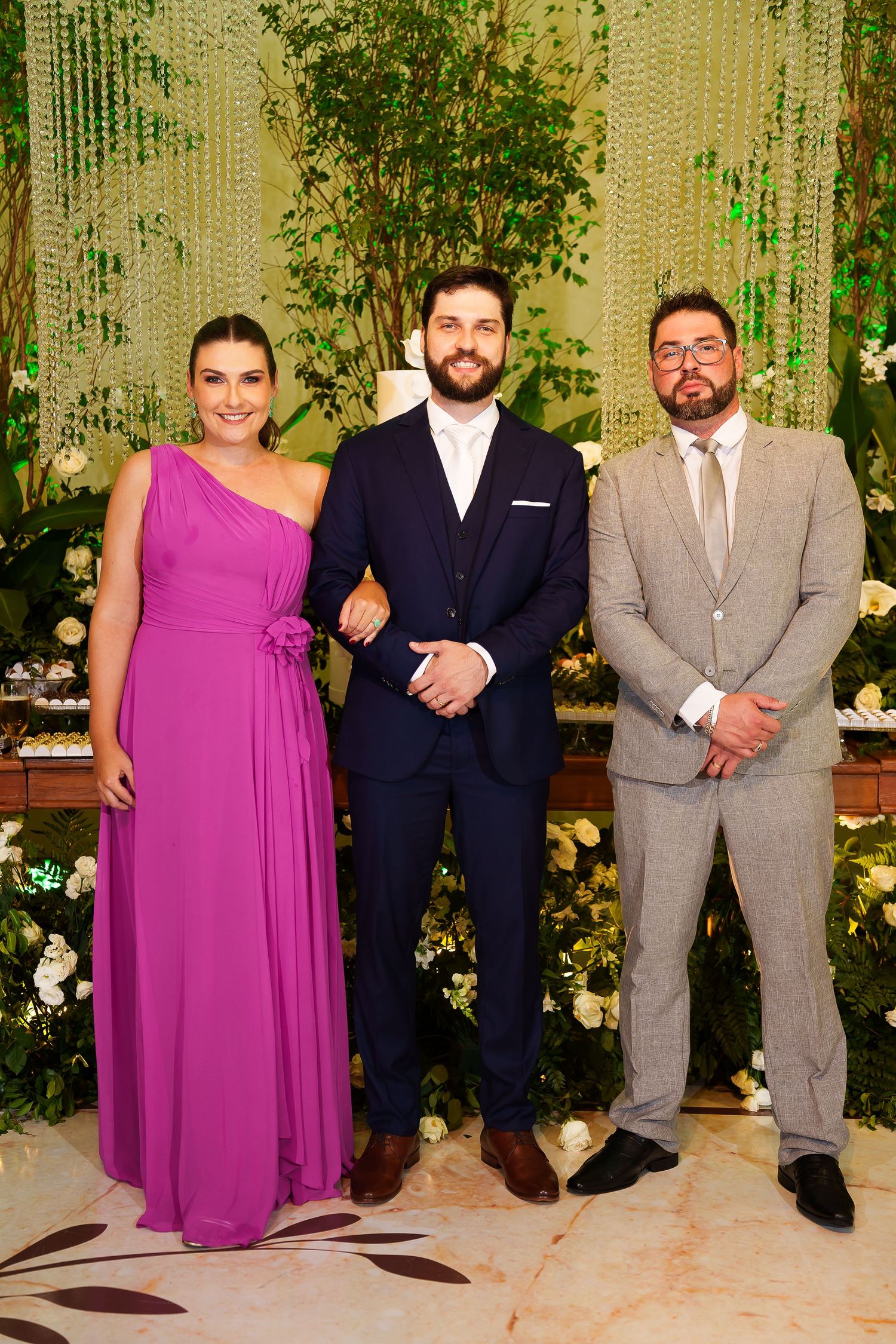 Three elegantly dressed guests pose together at a formal event, woman in magenta gown flanked by two men in suits.