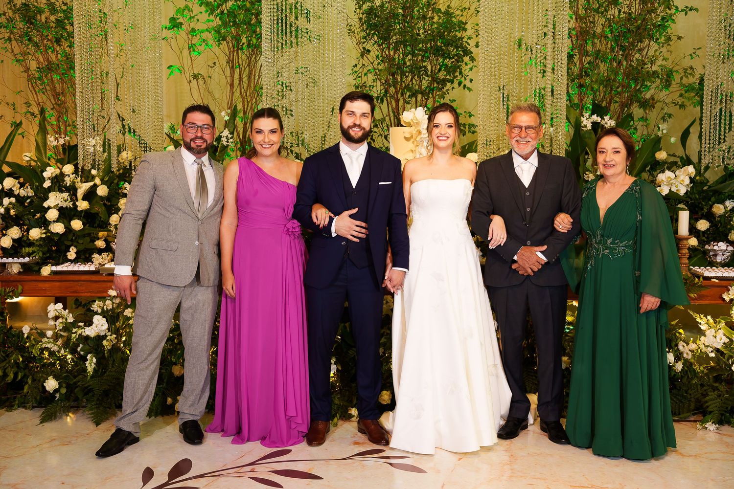 Wedding party of six posing together, bride in white gown, guests in formal attire, elegant floral backdrop.