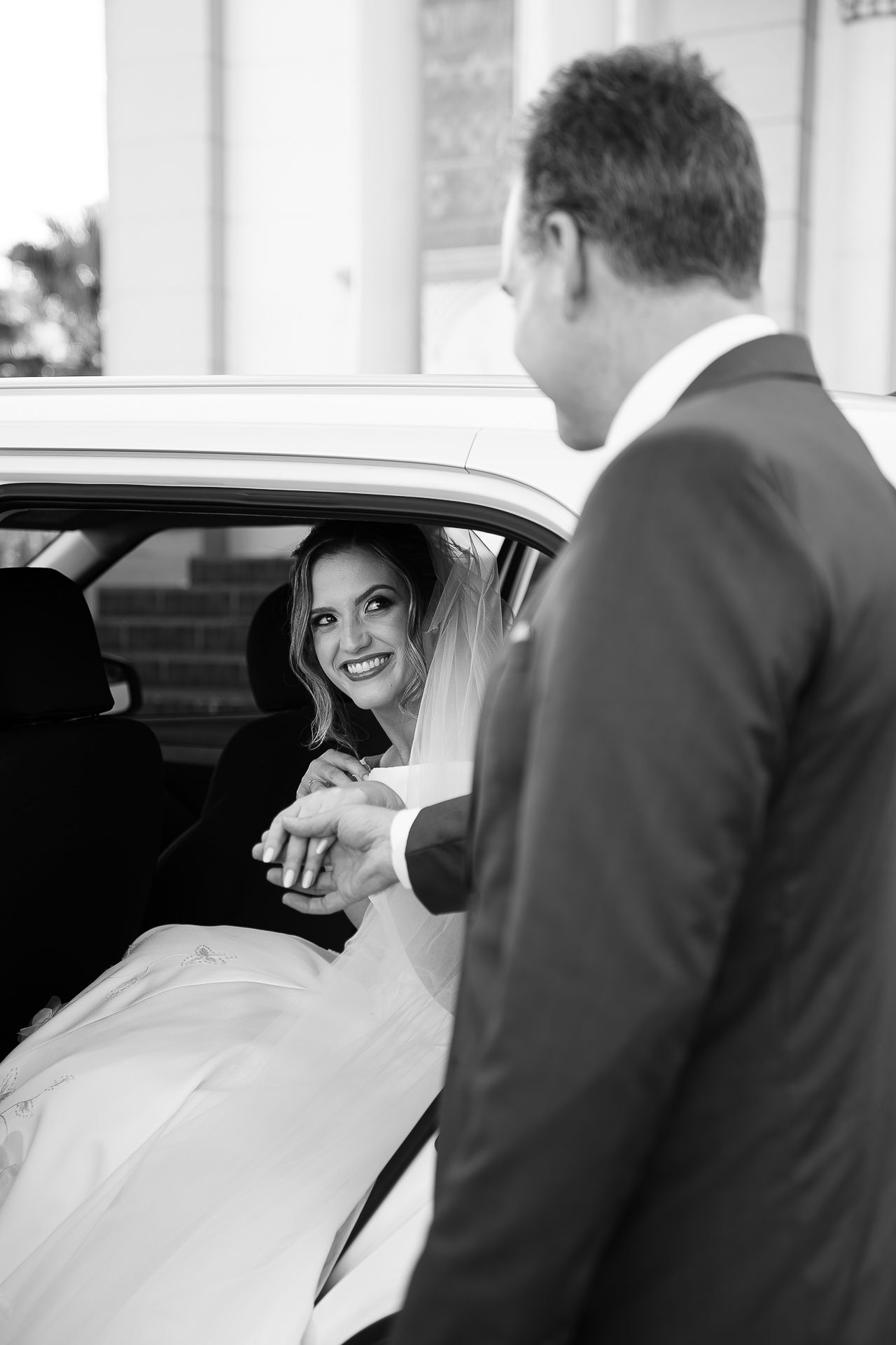 Black and white photo of a smiling bride in a wedding dress holding hands with a groom in a suit beside a car.